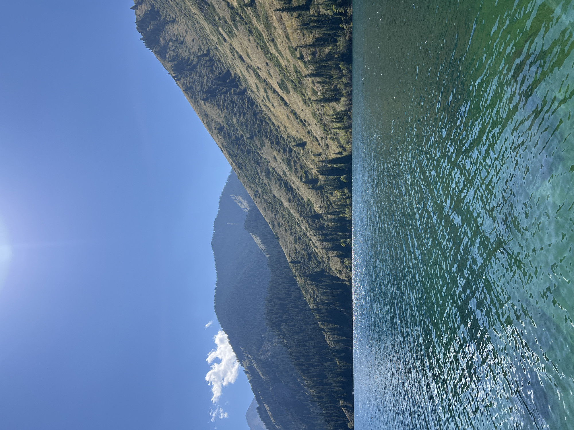 The image depicts a serene natural landscape of a lake bordered by mountains covered in coniferous trees and dry grass. The sky is a clear, bright blue with a sun flare visible in the upper left corner. The lake's water is a vibrant turquoise with ripples reflecting the sunlight. The mountains on the right side of the frame are a mix of grassy slopes and scattered evergreen trees, with a darker, densely forested mountain range in the background on the left. The overall scene suggests a peaceful outdoor setting, likely in a mountainous region. There are no people or discernible man-made structures in the immediate view.