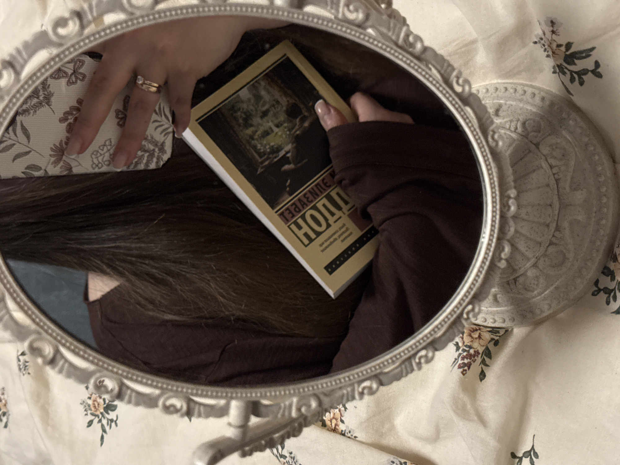The image depicts a close-up, reflected view of an individual holding a book and a smartphone, captured through an ornate, oval-shaped tabletop mirror with a distressed light-colored frame. The mirror rests on a light-colored fabric featuring a repeating pattern of muted orange-pink flowers and green leaves.
**People:** Portions of one individual are visible in the reflection. Long, dark brown hair cascades down the center. A maroon long-sleeved garment covers the upper torso. The individual's right hand holds a book, with the thumb visible on the spine and fingers extending across the cover. The individual's left hand holds a smartphone, with the thumb and fingers partially visible. The ring finger of the left hand bears two gold-colored rings: a prominent engagement-style ring with a clear stone, and a simpler band beneath it. Fingernails on both visible hands are manicured with a light-colored tip, consistent with a French manicure.
**Subjects/Objects:**
* **Mirror:** A decorative, oval-shaped mirror with an intricate, possibly plaster or resin, frame finished in an off-white or distressed pale color. The frame features scrollwork and beaded detailing.
* **Book:** A paperback or hardcover book with a yellowish spine and a dark cover. The title, reflected in the mirror, appears as "НОЛЛИВУД" (NOLLEWOOD or HOLLYWOOD in Cyrillic script). An illustration on the cover depicts a dark interior space with a view through an opening to green foliage.
* **Smartphone:** A smartphone is partially visible, held in the left hand. Its case is light-colored with a subtle pattern of dark botanical elements and butterflies.
* **Rings:** Two rings are worn on the left ring finger; one is a gold-colored band with a prominent clear stone, and the other is a plain gold-colored band.
**Actions/Interactions:** The individual is holding both a book and a smartphone. The composition suggests the image is a selfie taken by the individual using the smartphone, reflected in the mirror.
**Environment:** The immediate environment consists of the floral-patterned fabric surface upon which the mirror is placed, likely bedding or a tablecloth. No other definitive environmental features are discernible beyond the indistinct dark background reflected behind the individual's head.
**Location:** No discernible visual elements within the image provide information regarding a specific city, including Krasnodar, Russia. The items present are generic and could be found in various locations.