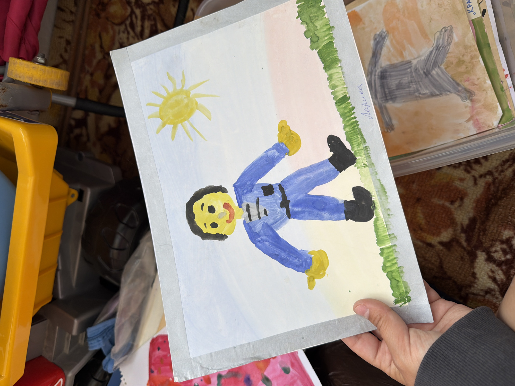 A child's watercolor painting of a person in a blue jumpsuit standing in a field is held up by a hand wearing a gray sweatshirt. The painting depicts a yellow sun in a pale blue sky, with a patch of green grass along the bottom. The figure has a yellow face with black hair, a red mouth, and black eyes. The jumpsuit has black details and a black belt. A name, "Лариса", is written in cursive script on the lower right side of the painting. The painting is placed on top of other children's artwork within a clear plastic container. The background suggests an indoor environment with a patterned rug and part of a yellow and red toy in the upper left corner.