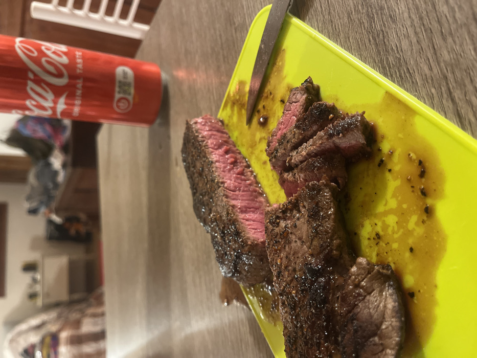 The scene depicts a domestic setting in Monsummano Terme, Italy, focusing on a meal prepared on a light wooden table. In the foreground, a vibrant lime-green plastic cutting board holds two cuts of cooked steak. One long, thick steak piece is severed, displaying a dark, seared exterior crust and a medium-rare, pink interior. Adjacent to it, another steak portion is partially sliced into thinner pieces, revealing a similar pink internal doneness and dark crust. The cutting board is stained with dark brown meat juices and black seasoning particles. A metal knife with a dark handle rests on the right side of the cutting board. Behind the cutting board, a red Coca-Cola can stands upright. The background is blurred but reveals elements of an indoor room: a striped bed or sofa on the far left, a small white dresser, a larger dark wooden dresser with items on top, and a white slatted chair on the far right. No individuals are present.