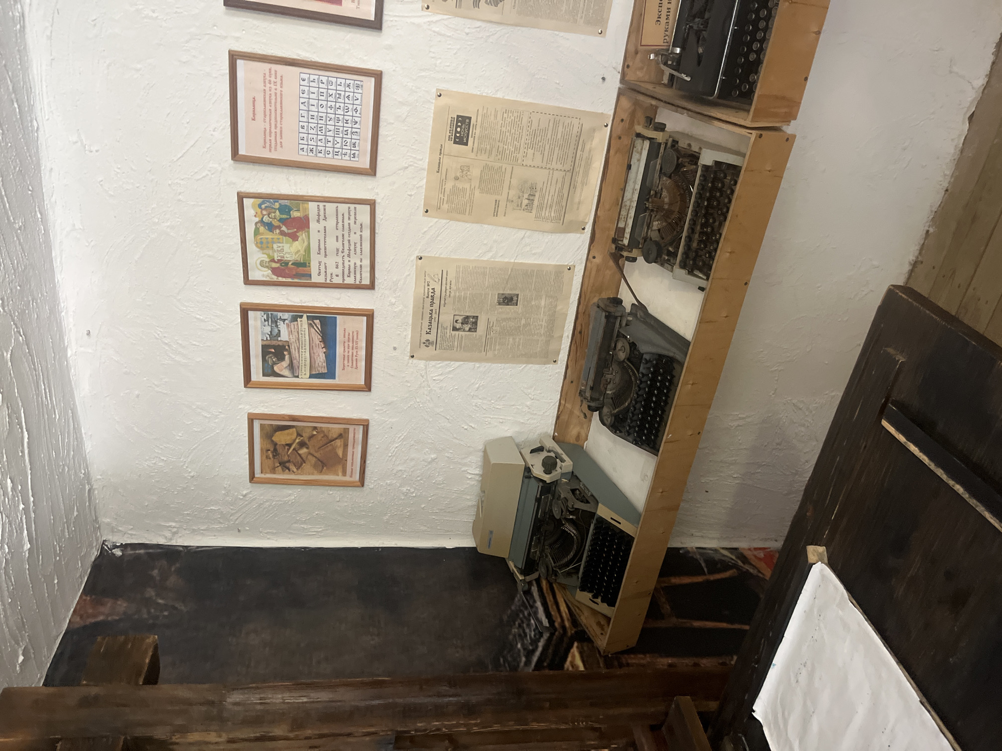 The image depicts an interior scene, likely within a museum or historical exhibit, in Taman', Russia. The wall is white and textured, with several framed prints and documents displayed. To the left, a large, dark mural is partially visible, covering a portion of the wall and extending to a wooden structural element.
In the foreground, a dark wooden table or counter is present. On the wall above a wooden shelf, four framed items are displayed. From left to right, the first frame contains a document with text and an illustration. The second frame shows a religious icon with figures. The third frame displays a grid of Cyrillic letters and text, labeled "Кириллица". The fourth frame also contains text and an image.
Below these frames, a wooden shelf holds several vintage typewriters. The first typewriter, on the left, is a light-colored machine with a grey base. Next to it are two older, darker-colored typewriters with visible keys and mechanisms. Further to the right, a black typewriter is partially visible in a wooden box. Two newspapers are pinned to the wall above the typewriters; the left one is titled "Казачья правда".
The overall environment suggests a historical display related to writing, journalism, or early Slavic literacy, possibly within the context of Cossack history given the newspaper title.