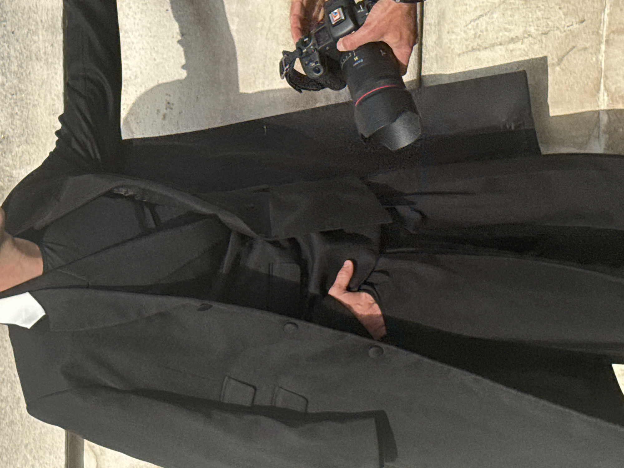 A person lying on the stone surface of a public area in London is being photographed. The person is wearing a black suit and white clerical collar. A photographer stands over the person, holding a professional DSLR camera with a large telephoto lens. The background consists of stone paving and architectural elements of what appears to be a public building or monument. A shadow suggests the presence of another individual standing nearby.