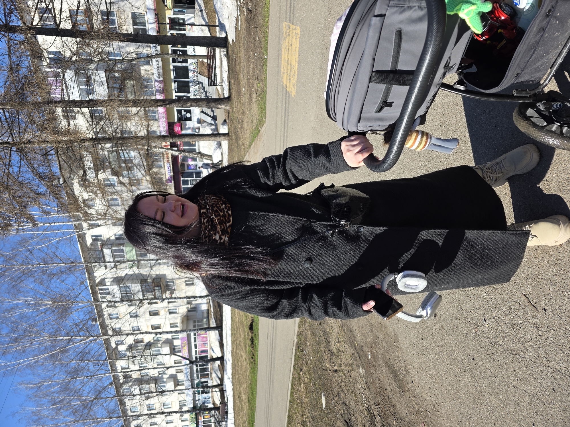 A woman with dark hair, wearing a long black coat and a leopard-print scarf, pushes a gray stroller on a sunny day in the city of Chaikovsky, Russia. She holds a black smartphone in her left hand and wears tan boots. White headphones rest against her coat. The stroller contains various items, including what appears to be a green toy and a red toy. Buildings and bare trees line the street. Patches of snow are visible on the grassy areas. A yellow stripe marks the pavement.