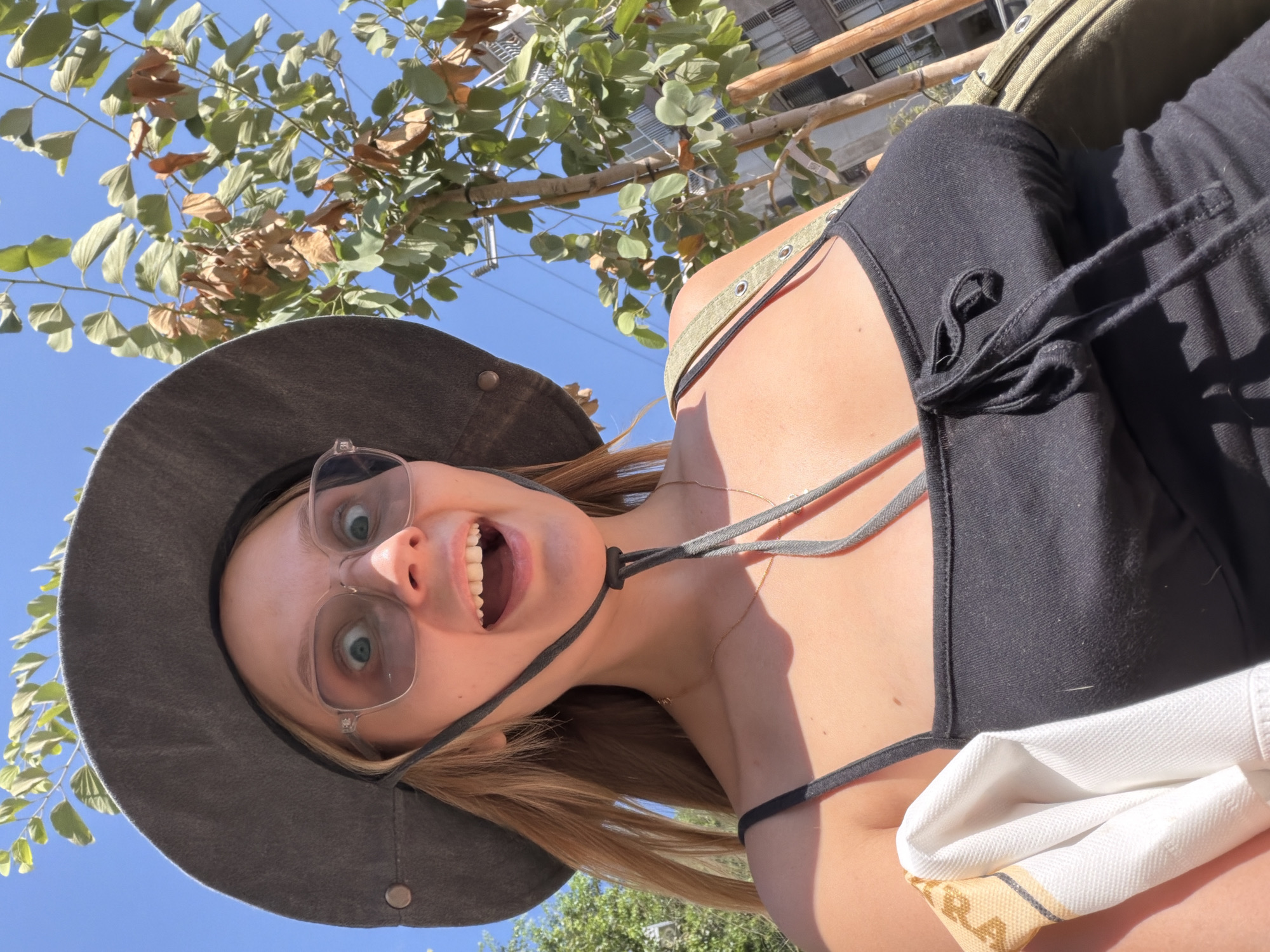 The media file is a selfie taken outdoors during the day, likely in Tel Aviv, Israel. The subject is a young woman wearing a wide-brimmed hat and a black top. She is looking directly at the camera with an open mouth and wide eyes, appearing excited or surprised. Her hair is blonde and falls around her face. She is wearing glasses with clear frames. The background shows a clear blue sky and green tree branches with some dried leaves, suggesting a warm, possibly late summer or early autumn day. The top she is wearing has thin straps with metal eyelets. A white cloth with a yellow and black pattern is visible resting on her lap. The image is oriented sideways.