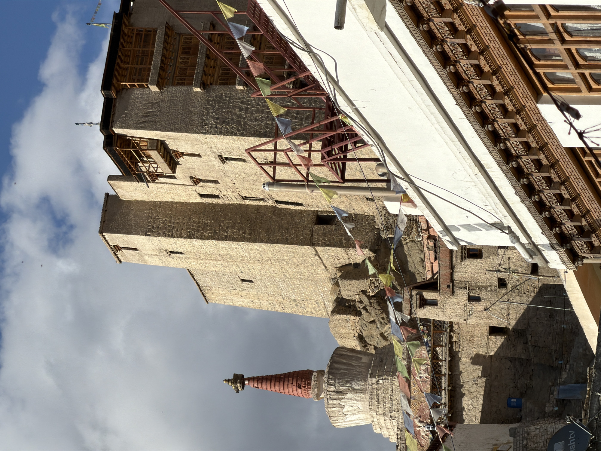 The image shows a view of the Leh Palace, a former royal palace in Leh, Ladakh, India. The palace is a tall, multi-storied structure built of stone and mud brick. It is a prominent landmark in the city, and its construction dates back to the 17th century. The palace has undergone several renovations and additions over the years, reflecting the changing architectural styles and needs of the region. In the foreground, there are prayer flags strung across a white wall. The prayer flags are a common sight in Buddhist regions of India and are believed to carry prayers and blessings on the wind. The sky is partly cloudy, with a few birds flying in the distance. The overall scene depicts a blend of historical architecture and cultural elements, characteristic of Leh, a city known for its rich heritage and Buddhist traditions.