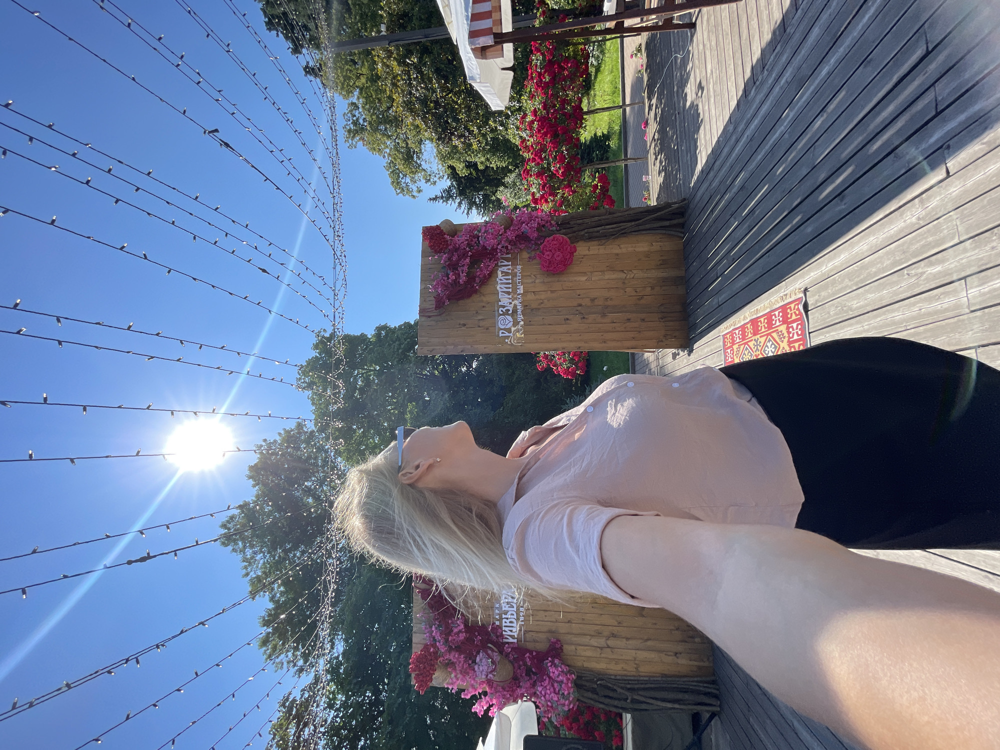 The image shows a blonde woman wearing a pink blouse and black skirt, posing for a selfie outdoors. She is wearing sunglasses and looking to her right. In the background, there are two wooden structures decorated with pink and red flowers, and trees. String lights are visible across the top of the image, and the sun is shining brightly. The woman is standing on a wooden deck with a patterned rug. The scene appears to be at "Riviera Park" in Sochi, Russia, as indicated by text on the wooden structures.