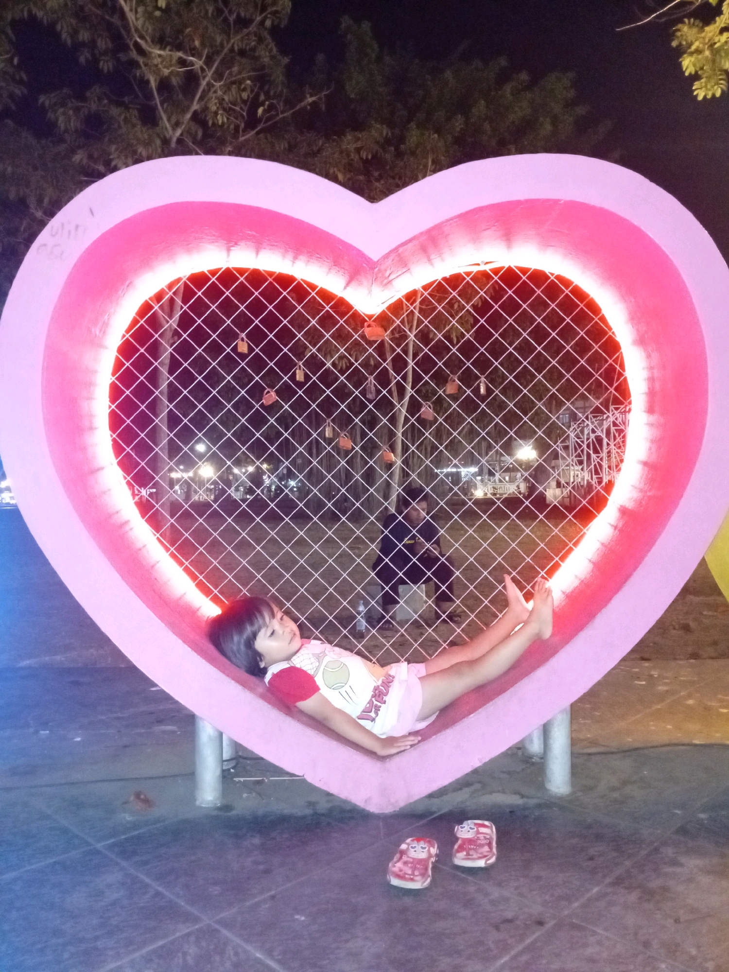 A young child is lying inside a large, pink heart-shaped structure illuminated by pink lights at night. The child is wearing a red and white shirt and pink shorts. The heart structure has a mesh fence inside it, and beyond the fence, a man is visible sitting and looking at his phone. In front of the heart structure, on the ground, are a pair of pink child-sized sandals with cartoon characters on them. The ground appears to be paved with tiles. The scene is likely located in a public space within the city of Purworejo, Indonesia, possibly a park or a tourist attraction decorated for evening viewing. Trees and distant lights are visible in the background.
