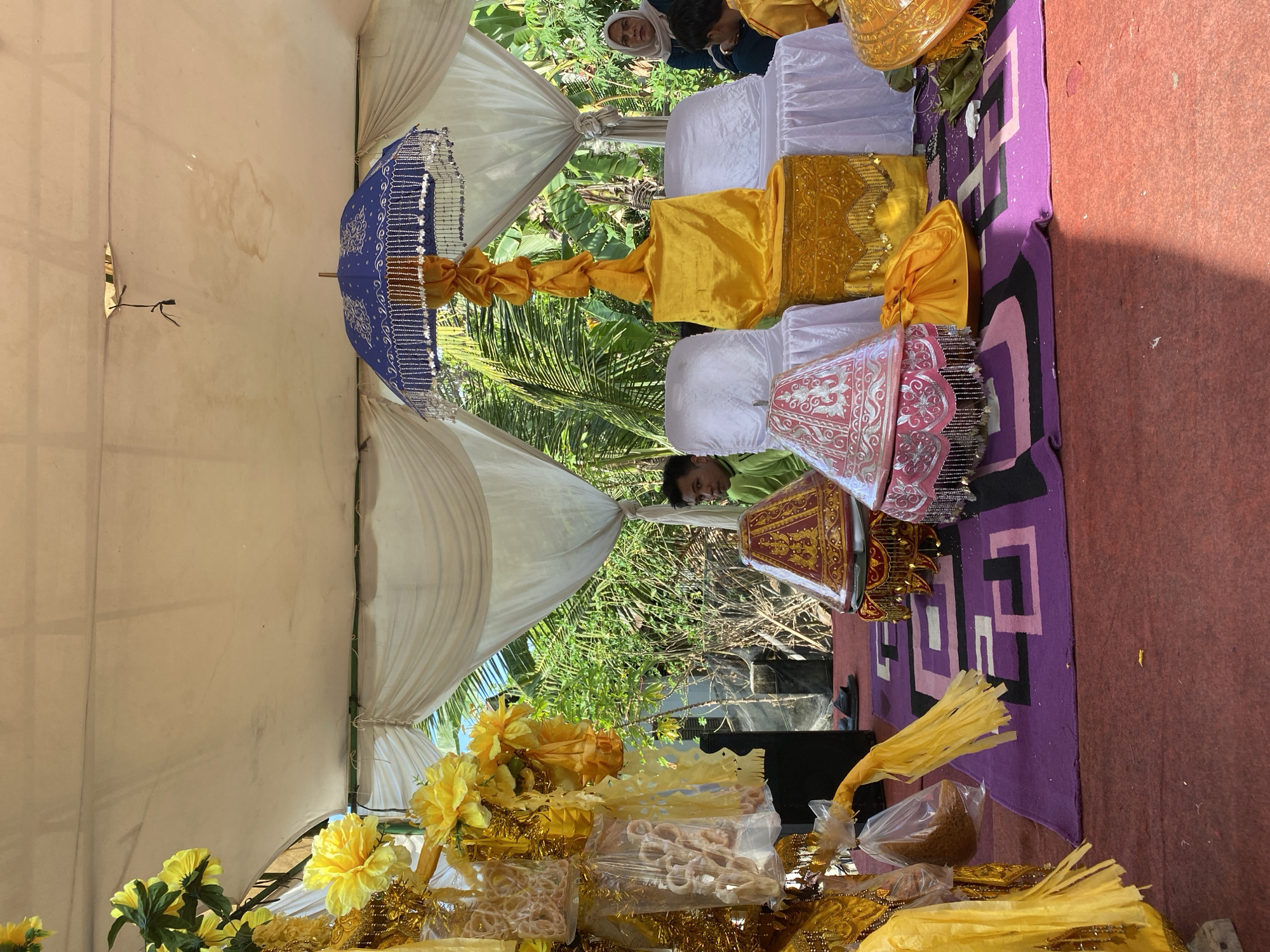 An outdoor event is being held under a white tent in the city of Latong, Indonesia. The area is decorated with yellow and gold fabric. There are several people present, including a man in a green shirt and a woman in a headscarf. In the foreground, there are ornate decorative items, including a blue umbrella with silver trim, and two conical objects covered in red and pink fabric with intricate patterns and beaded fringe. The ground is covered with a red carpet, and a purple rug with geometric patterns is placed on top of it. There are also bags of what appear to be snacks hanging from a decorative stand on the left side of the image. The background shows lush green foliage, suggesting a tropical environment.