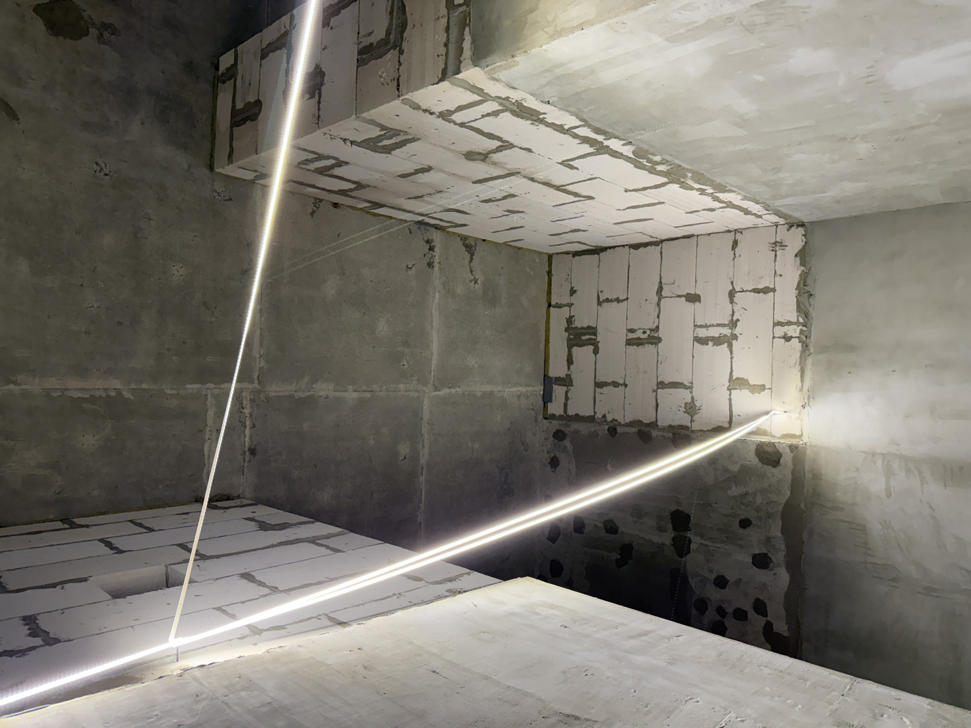The image depicts the interior of an unfinished building, likely under construction. The walls are made of concrete and light-colored cinder blocks. Several thin, illuminated LED light strips are visible, tracing geometric patterns across the ceiling and walls. The lighting suggests an artificial, possibly contemporary, aesthetic being installed. The overall environment is raw and industrial, with visible construction materials and textures. There are no people present in the scene. The location in Moscow, Russia, is not determinable from the visual information provided.