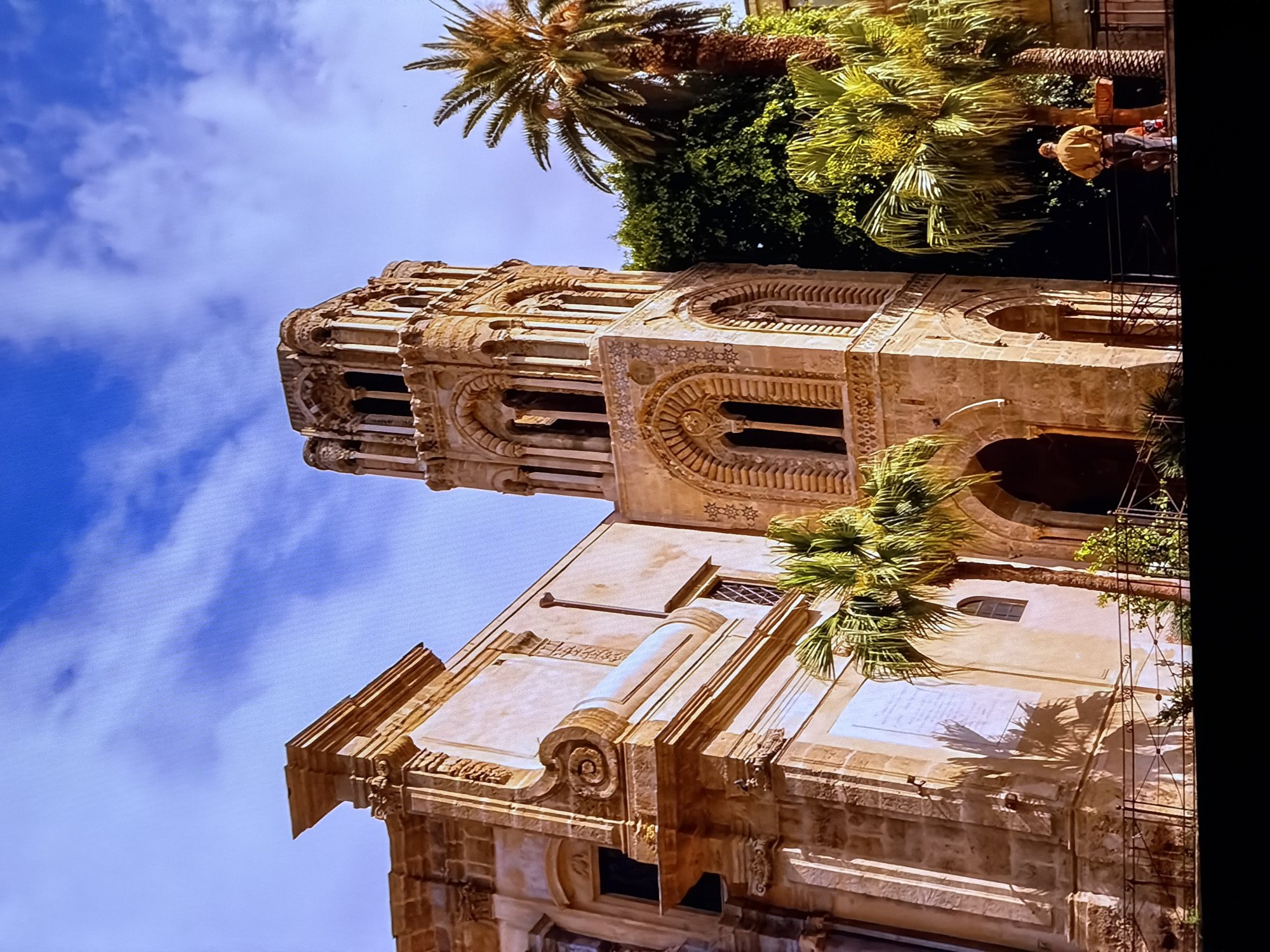 This image shows an exterior view of a historic stone building with a bell tower and palm trees in the foreground. The architecture appears to be a blend of Romanesque and Islamic styles, with intricate carvings and arched windows. The sky is a vibrant blue with scattered clouds. A person in a yellow shirt and jeans is standing near a tree, facing away from the camera. The scene is well-lit, suggesting it is daytime. There are no visible elements that indicate the location in the city of Chirchik, Uzbekistan.