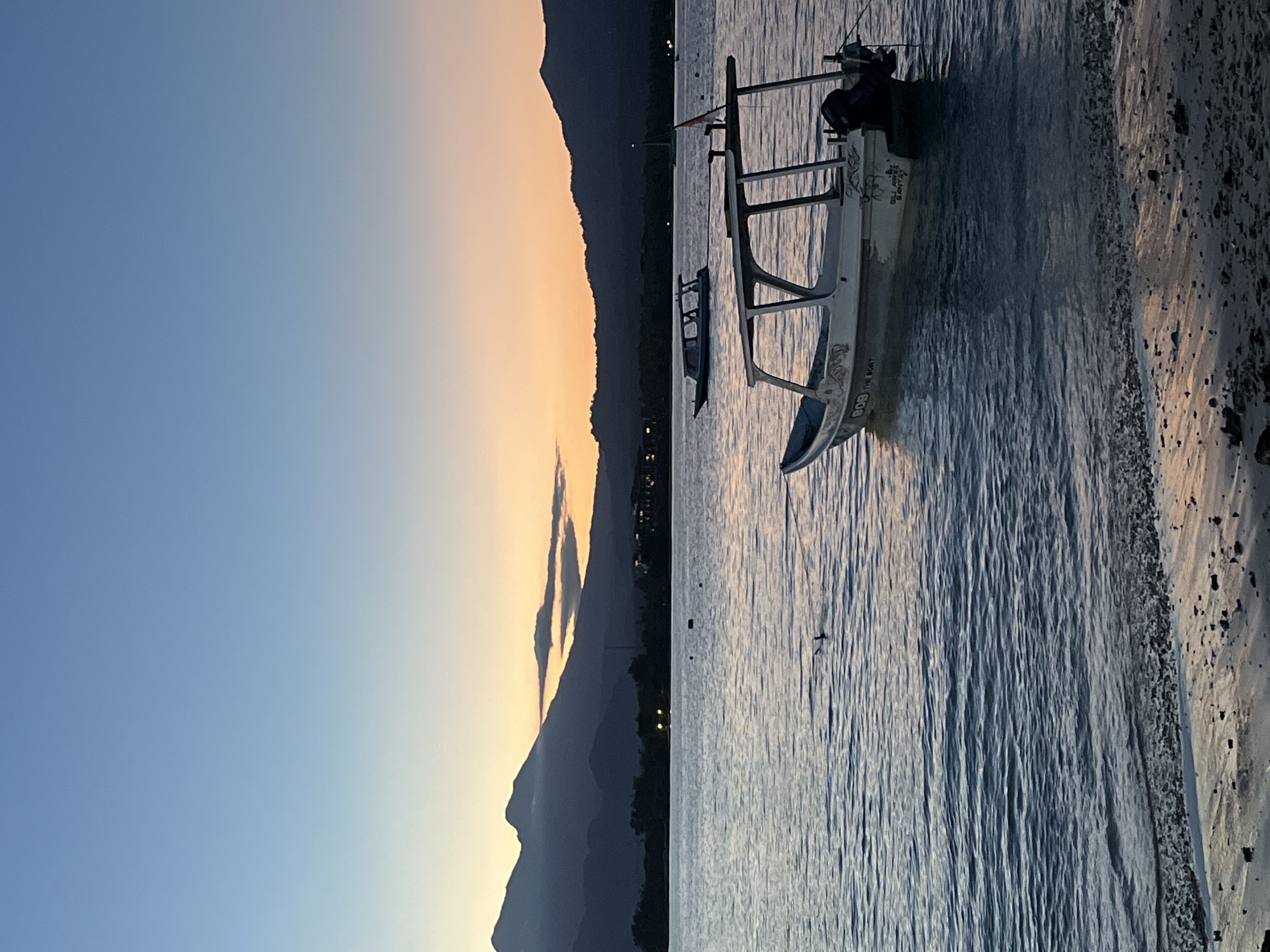 A landscape photograph, taken in an unknown city in Indonesia, presents a seascape at dawn or dusk, with a boat anchored near the shore. The photograph is framed with the horizon running across the center. The left half of the image is dominated by the sky, which graduates from a deep blue to a pale orange, suggesting either sunrise or sunset. In the background is the silhouette of a mountain range, with the rising or setting sun casting a golden glow along its ridges. The right half of the image is occupied by the sea, which appears calm with small ripples. A traditional Indonesian fishing boat, possibly a Jukung, is docked close to the shore. The hull of the boat is white with visible inscriptions on it. The shallow shoreline is visible at the edge of the water, a mixture of sand and small rocks. No people are clearly visible, though there might be a small figure onboard the boat. Small lights twinkle on land by the mountain, likely from houses.