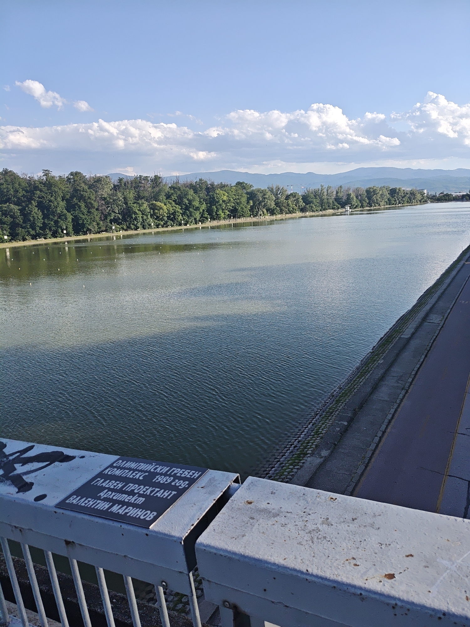 An elevated, wide-angle shot captures a serene river scene under a partly cloudy blue sky. The river dominates the frame, its surface a dappled mix of green and blue hues, reflecting the sky and surrounding trees. A line of lush green trees forms a dense border along the far bank, with distant, hazy mountains providing a backdrop. To the right, a paved pathway runs alongside the river, separated from it by a stone embankment. In the foreground, the railing of a bridge or elevated walkway is visible, adorned with a metal plaque inscribed with text in Cyrillic. The plaque reads: "ОЛИМПИЙСКИ ГРЕБЕН КОМПЛЕКС 1989 ГОД. ГЛАВЕН ПРОЕКТАНТ Архитект ВАЛЕНТИН МАРИНОВ", which translates to "OLYMPIC ROWING COMPLEX 1989 YEAR. CHIEF DESIGNER Architect VALENTIN MARINOV". The railing itself shows signs of wear, with some graffiti and rust spots. The overall atmosphere is peaceful and expansive.