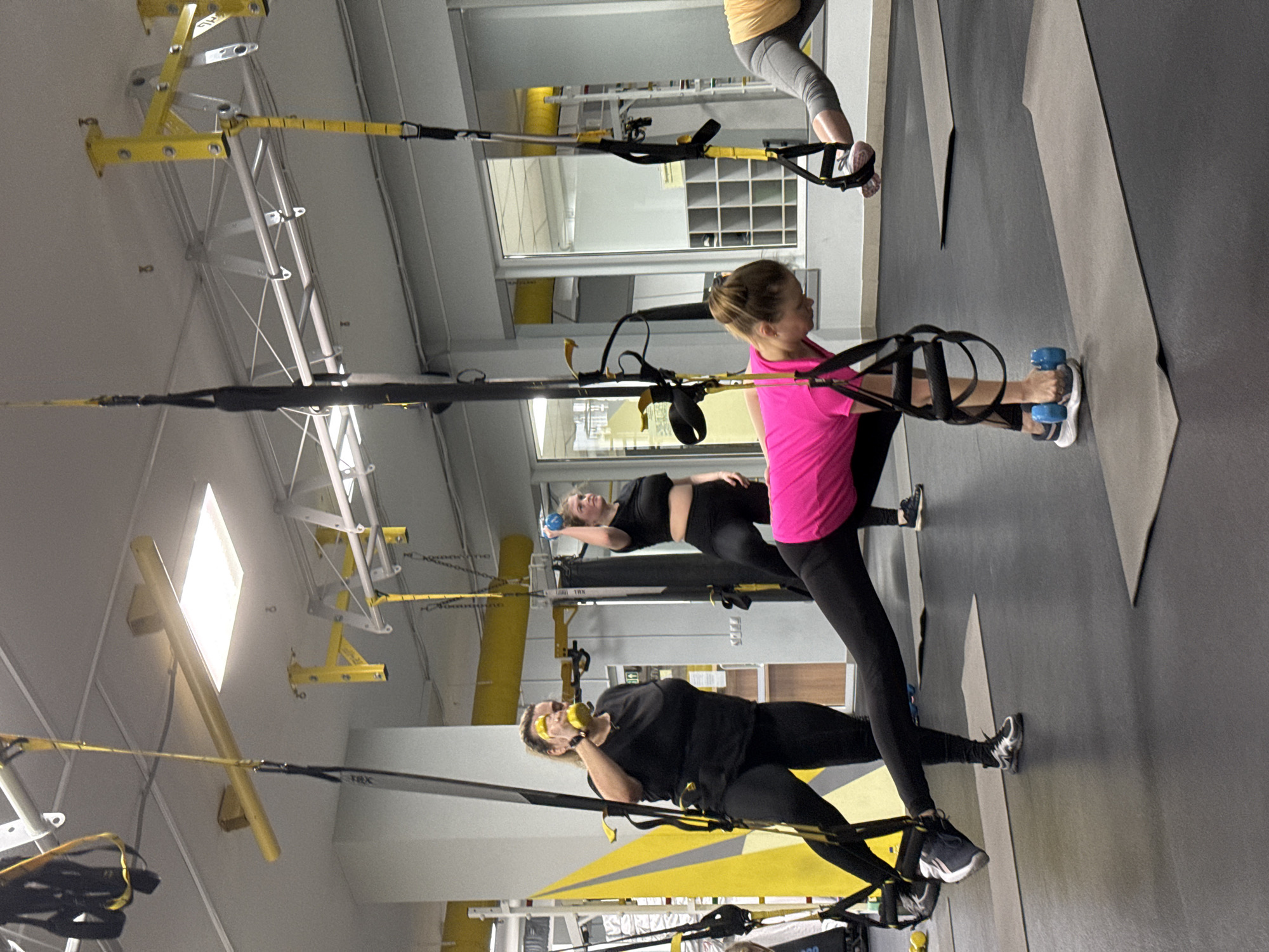 Three women are exercising with TRX suspension trainers in what appears to be a gym. The gym has a gray floor and a white ceiling with yellow support structures for the suspension trainers. The woman closest to the viewer wears a black shirt and black leggings, and is using the TRX straps. The next woman wears a black shirt and black leggings. She is holding a blue dumbbell. The third woman wears a bright pink shirt and black leggings. She is also using TRX straps while holding a blue dumbbell. The location is possibly Chaikovsky, Russia.