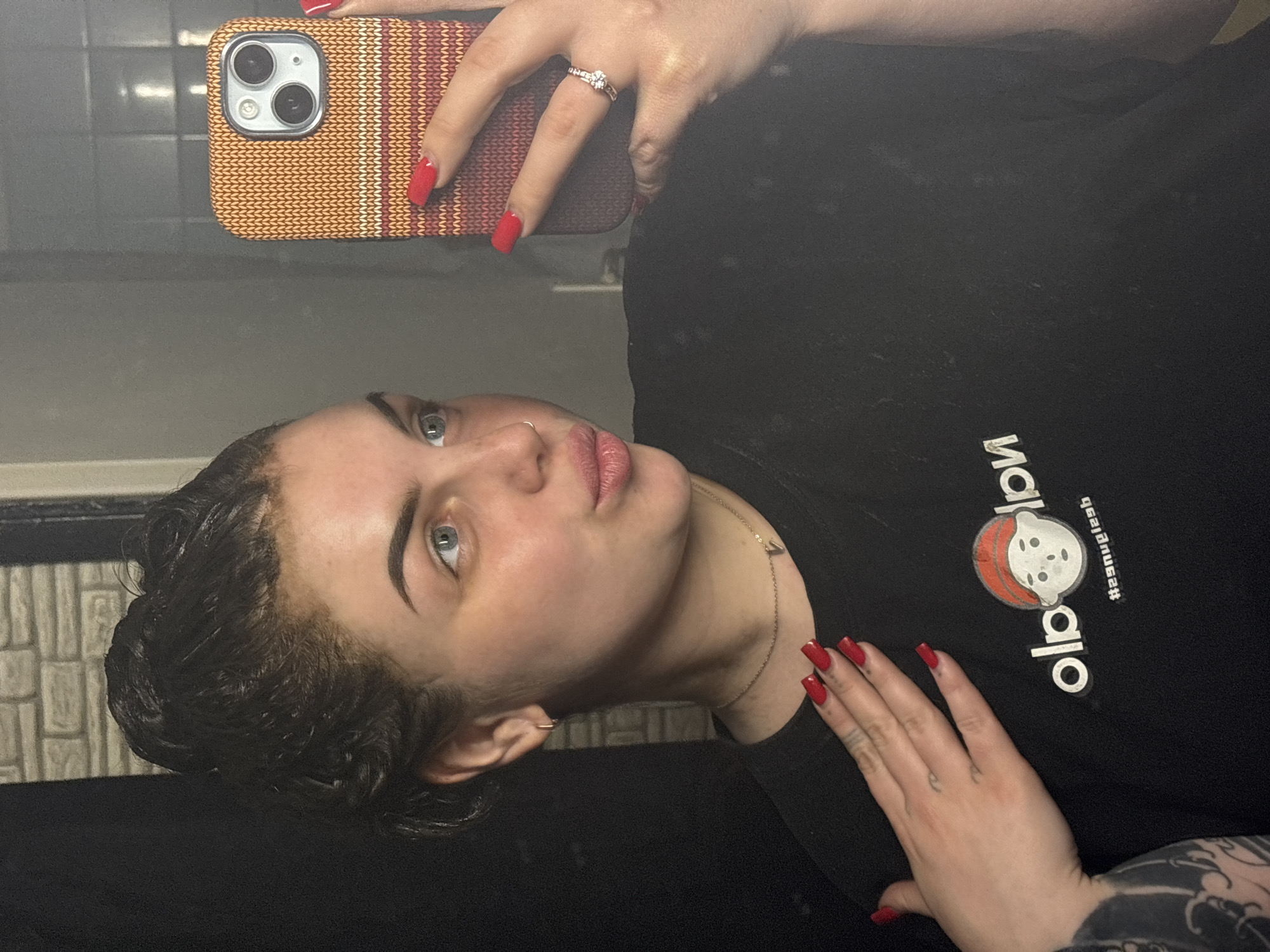 A person is taking a selfie in what appears to be a bathroom. The person has wet, dark hair, light eyes, and is wearing a black t-shirt with a graphic print that includes the reversed word "old". They are holding a smartphone with a knitted-looking case, displaying red fingernails and a ring on their finger. The background includes tiled walls and a mirror. The location of Chornomorskoe, Ukraine is not directly evident in the image.