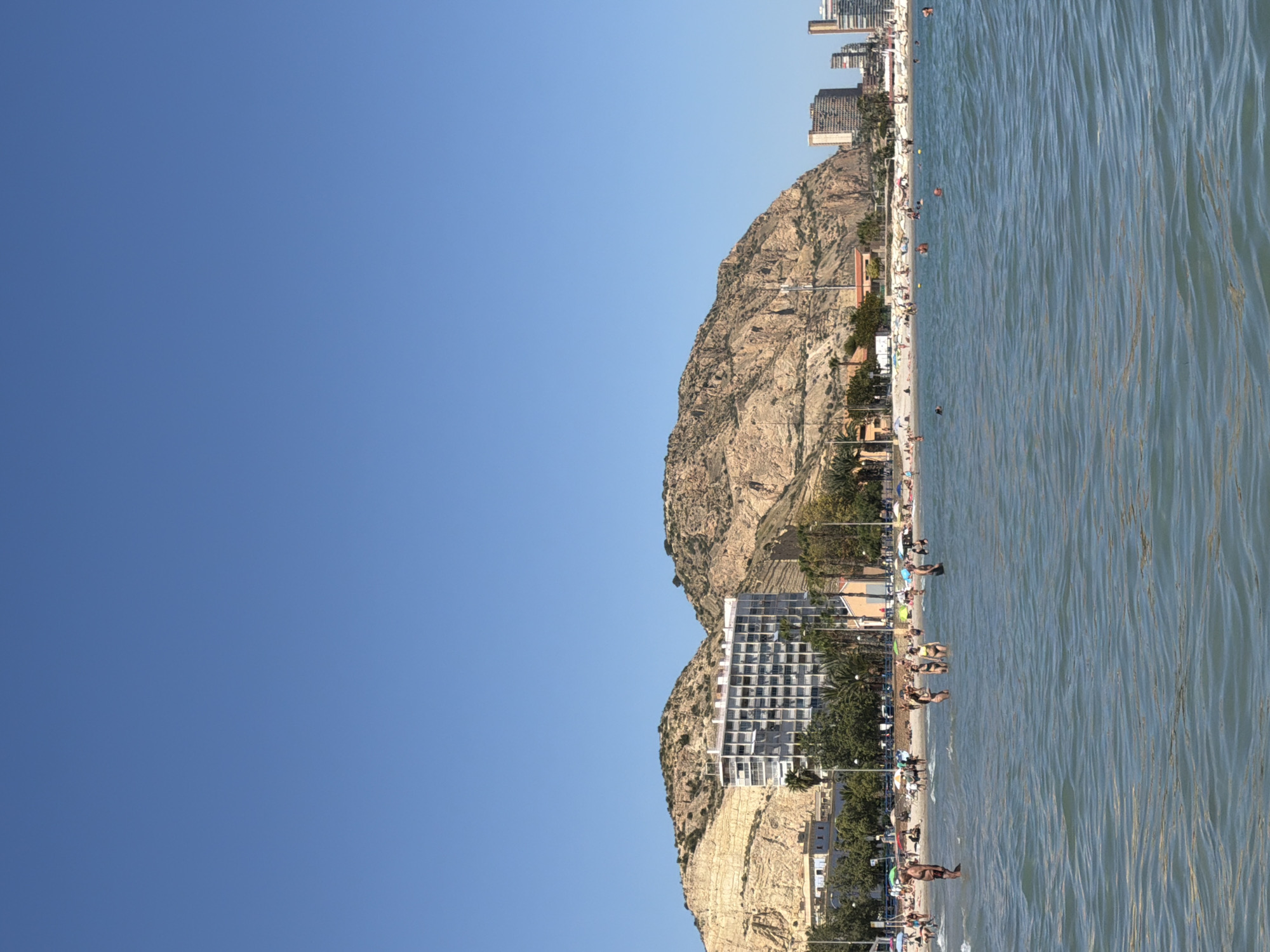 The image depicts a coastal scene in an unidentified city. It showcases a beach along the shoreline, featuring a number of people who seem to be enjoying recreational activities such as swimming and sunbathing. There is a notable, rocky landmass in the background, with buildings interspersed among the landscape, and further back a row of tall buildings. The right side of the image captures the sea, exhibiting a rippled texture and visible water movement. The sky is clear and blue on the left.