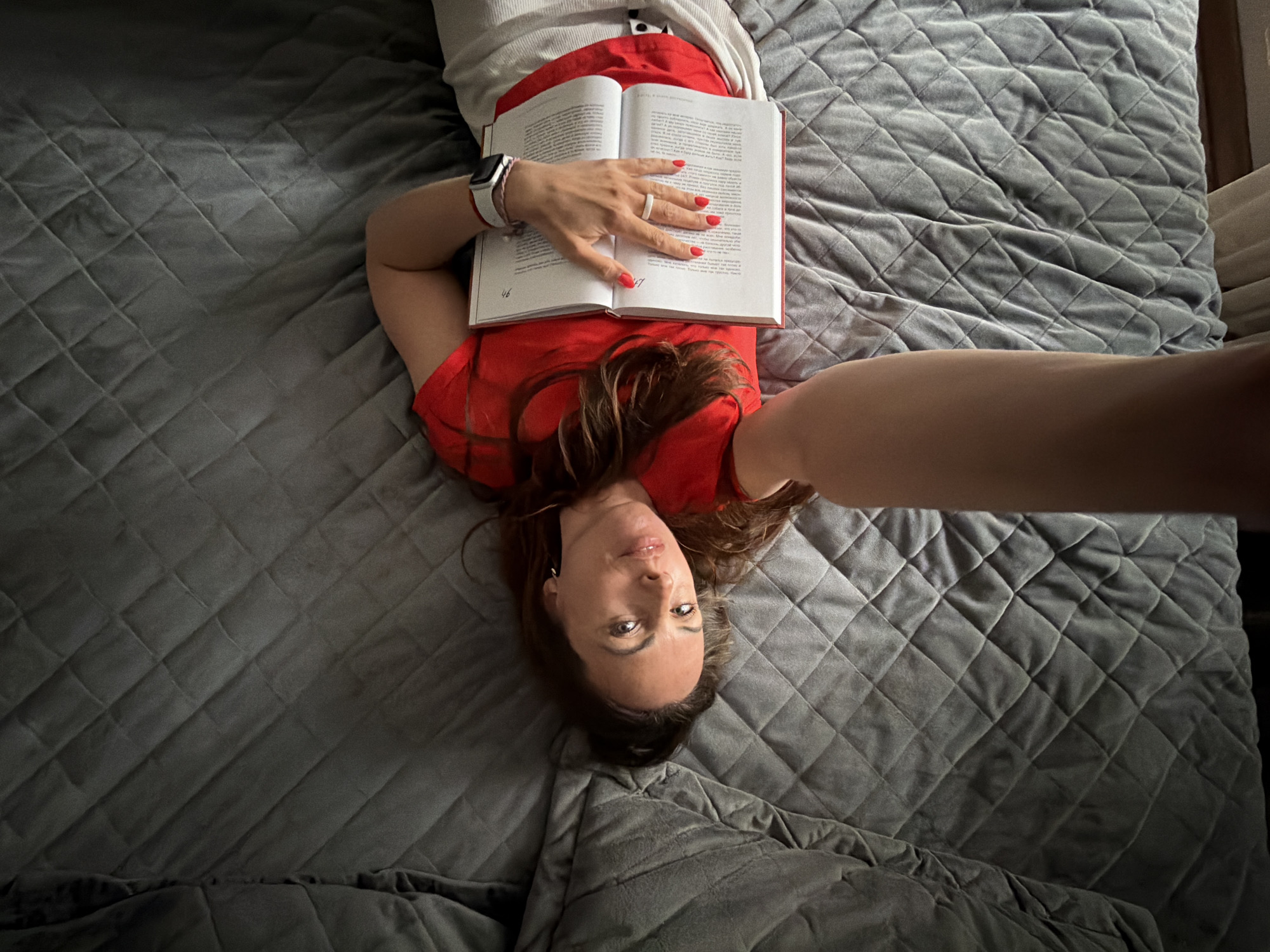 A woman lies on a gray quilted bed, holding open a book with red covers, pages 46 and 47 visible. She wears a red top and a white bracelet watch. Her nails are painted red. The photograph appears to be a selfie, taken at arm's length. There is no indication of Birobidzhan, Russia.