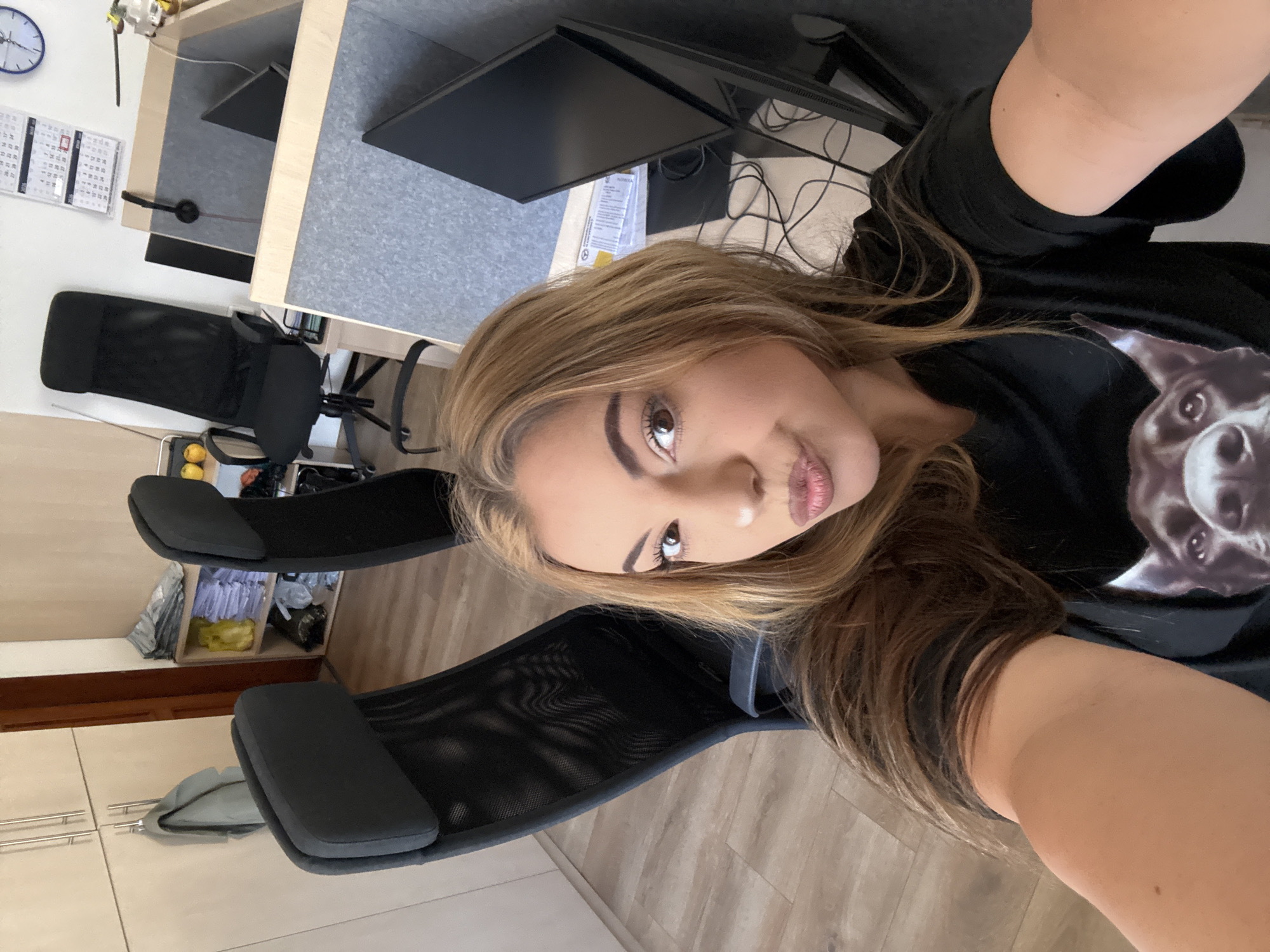 The image is a selfie of a young woman with light brown hair and fair skin. She is wearing a black t-shirt with a graphic print of a dog. She is sitting in a black mesh office chair. Behind her are office desks with computer monitors. In the background, there are office cabinets and a wall calendar. There is no information available to definitively ascertain the location in the city of Sofia, Bulgaria from the image.