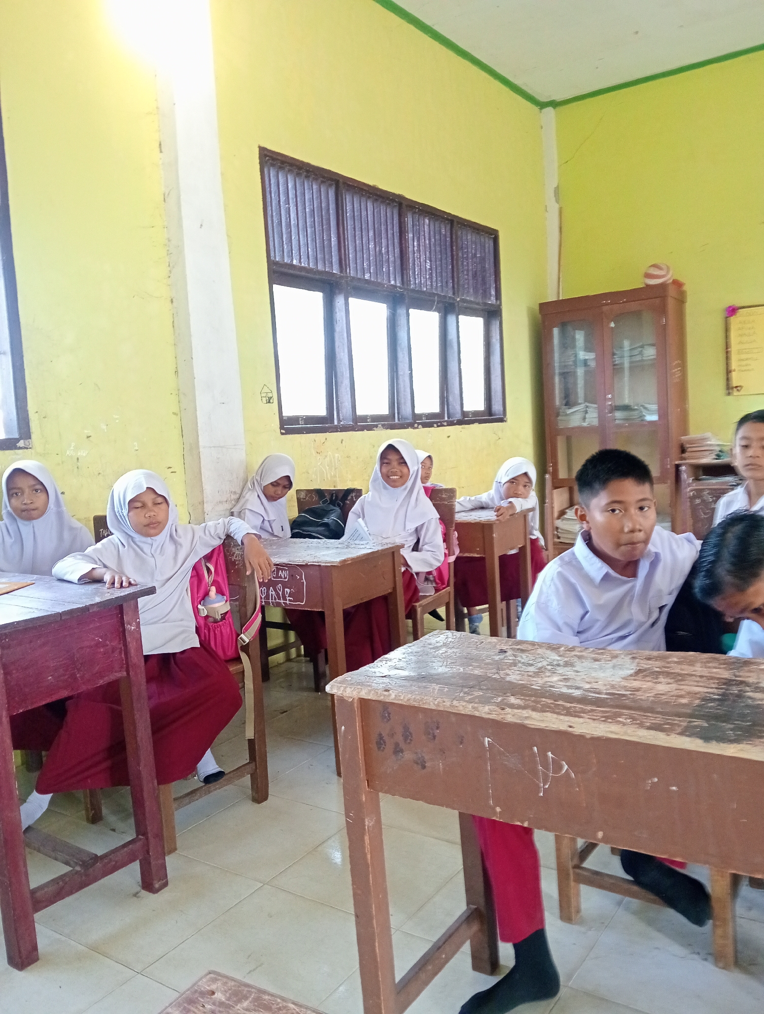 The image depicts a classroom scene in Keudekrueng, Indonesia, where several students are seated at wooden desks. The room has light yellow walls and a ceiling with green trim. A large window with dark vertical bars is visible on the left side of the image, and another window is present on the right. A wooden cabinet with glass doors stands against the back wall. The students are dressed in school uniforms, with the girls wearing white hijabs and maroon skirts, and the boys wearing white shirts. Some students are looking at the camera, while others appear to be engaged in activities at their desks. The desks themselves are made of wood, with some showing signs of wear and graffiti. A pink backpack is visible on the back of one student's chair. The floor is tiled with a light-colored pattern. The overall impression is that of a typical Indonesian primary school classroom.