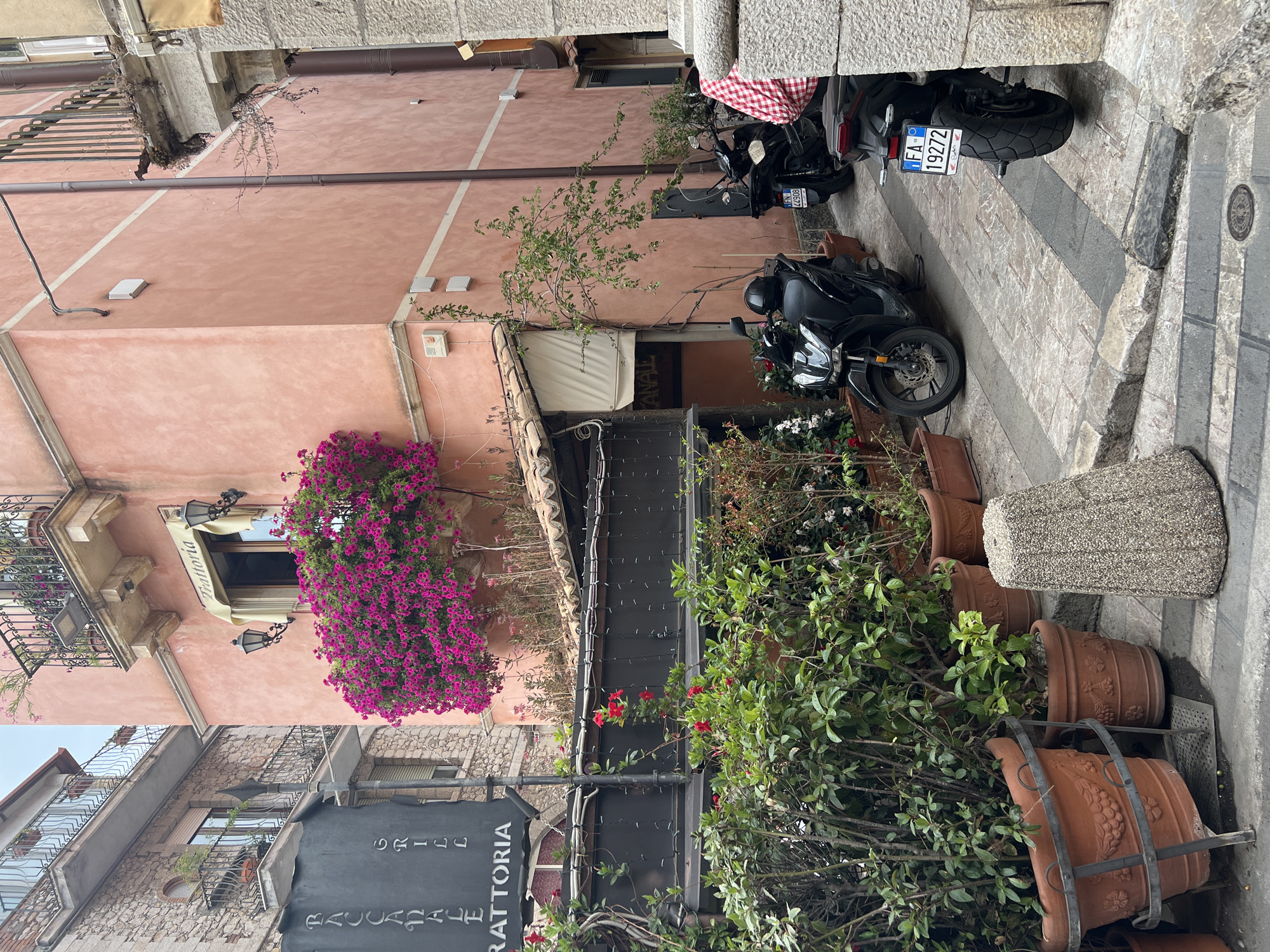 The image depicts a narrow cobblestone alleyway in Taormina, Italy, flanked by buildings with a distinct Mediterranean architectural style. The building on the right is painted a warm, peachy-pink hue and features a balcony adorned with lush pink bougainvillea cascading over its railing. Two black scooters are parked in the alley. The building on the left is constructed of stone and has balconies with ornate iron railings. A dark gray sign with white lettering that reads "RACCANALE GRILL TRATTORIA" is prominently displayed. Greenery, including potted plants and vines, lines the lower section of the alley, creating a vibrant, natural contrast with the man-made structures. The overall atmosphere is one of charming, old-world Italian street life.