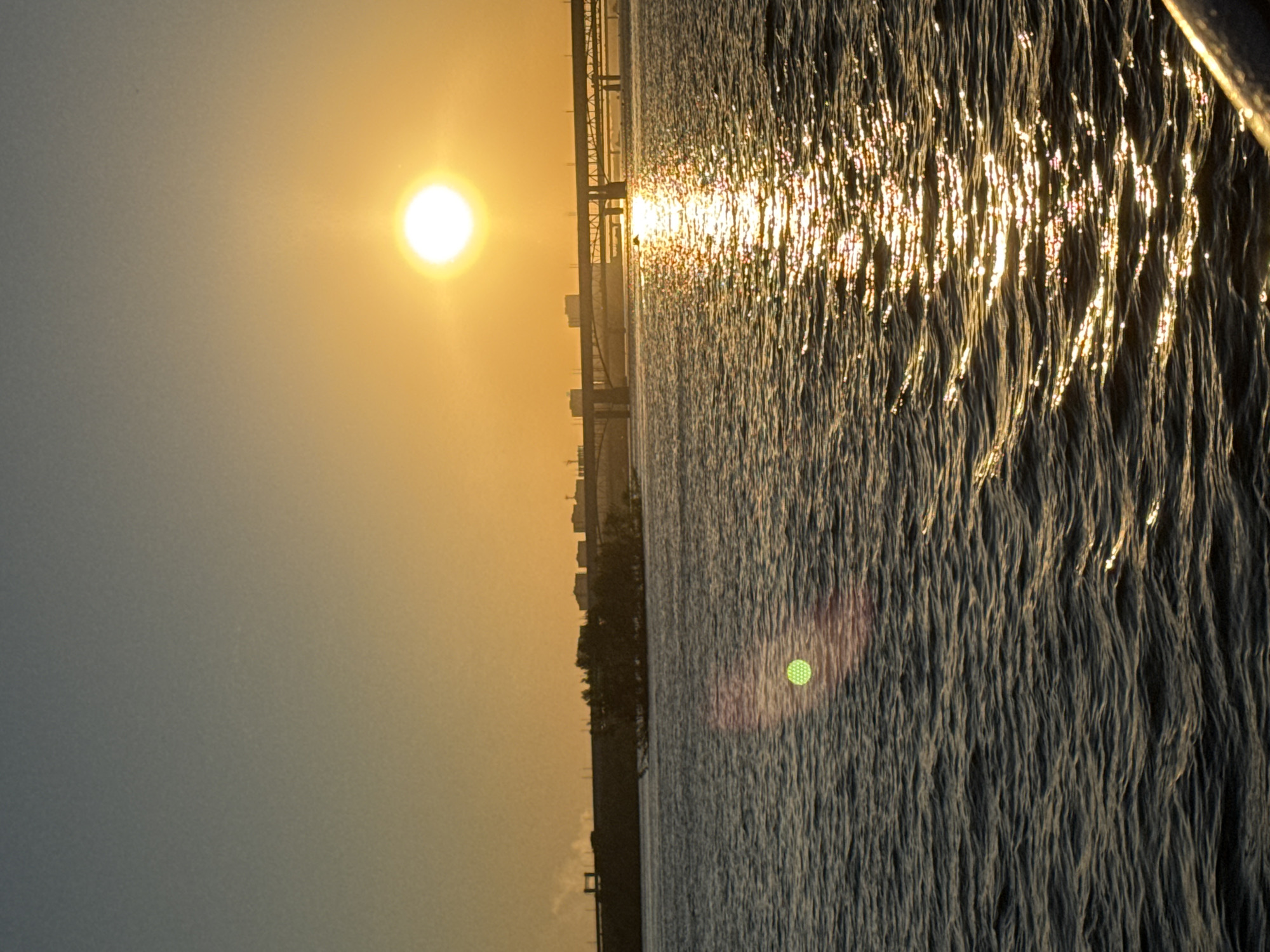 This image depicts the sunset over the Ob River in Novosibirsk, Russia. The sun, a bright, luminous orb, is positioned in the upper right quadrant of the sky, casting a warm golden glow across the scene. A prominent bridge, likely part of the Novosibirsk Metro Bridge or the Oktyabrsky Bridge, spans the river in the mid-ground, with its structure silhouetted against the radiant sky. On the distant bank, a city skyline with buildings is visible. The river's surface is rippled, reflecting the sunlight in a shimmering path that stretches towards the viewer. In the left mid-ground of the water, a green, hexagonal lens flare is present. The overall atmosphere is serene, with the setting sun illuminating the water and the city.