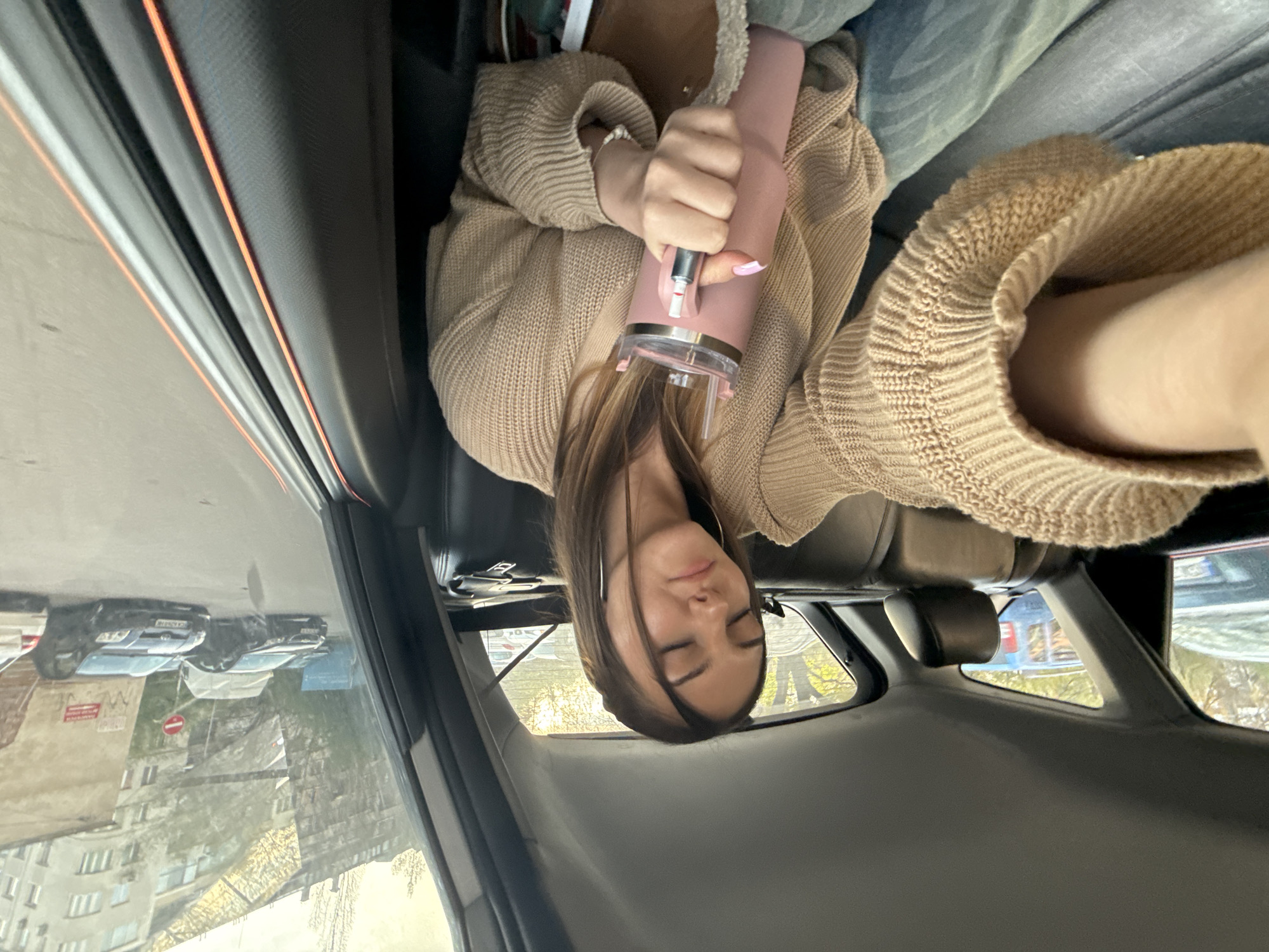 A young woman is sitting in the back seat of a car, holding a pink water bottle. She is wearing a tan-colored sweater and light blue jeans. The car's interior is visible, including the seats and windows. Outside the car, a street scene can be observed, showing buildings, cars, and foliage. Based on the visual characteristics, the location is likely Sofia, Bulgaria.