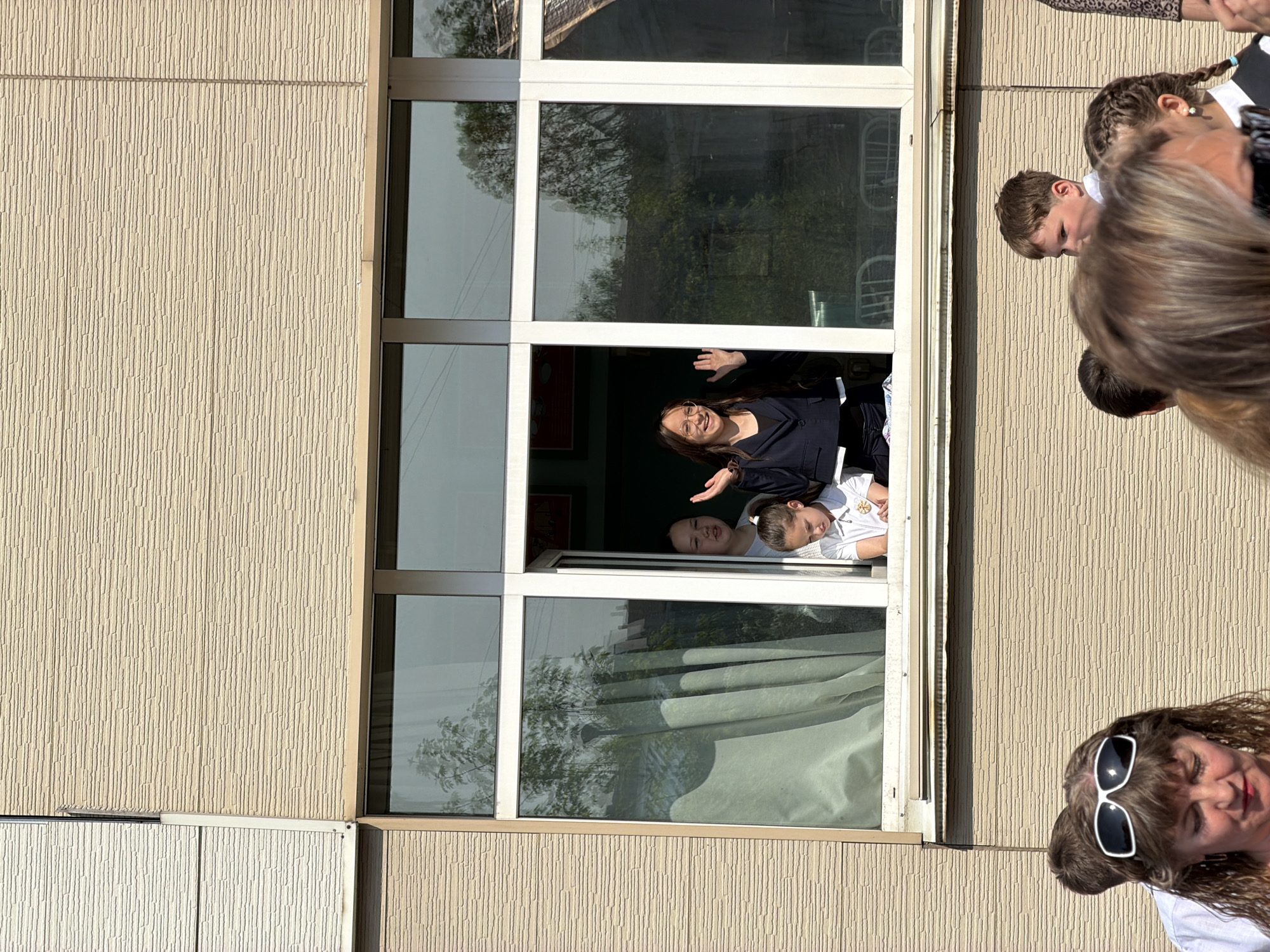 Outside a building with textured, beige walls and white window frames, a group of people is gathered. A window is open and three people are visible inside. A woman with long, brown hair wears glasses and a dark jacket; she smiles and waves. A younger person in a white top leans out the window. An additional person is visible in the window next to them. A woman with sunglasses is near the window, looking upwards. Other people are clustered near the window. Based on the information provided, this is taking place in Birobidzhan, Russia.