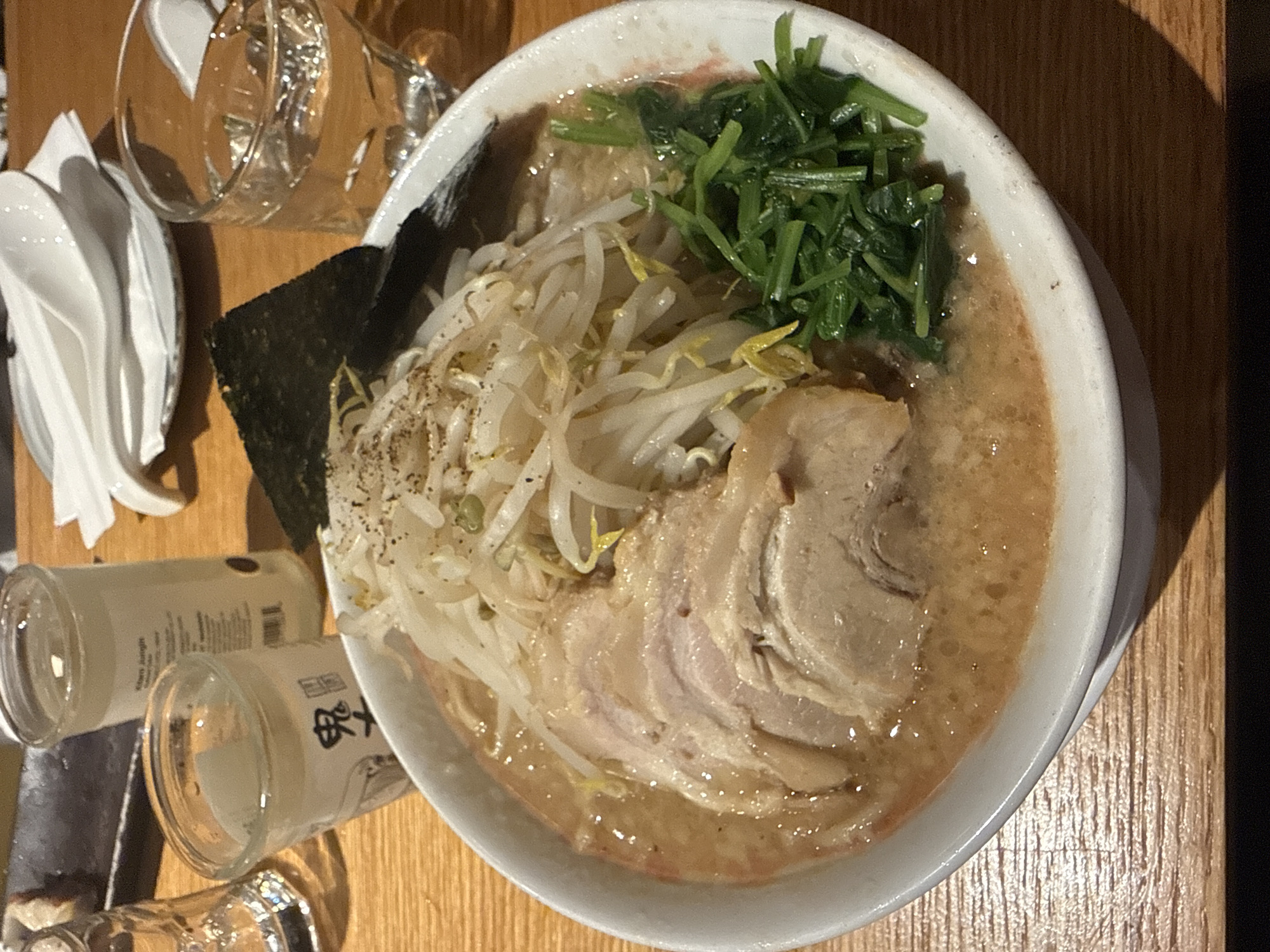 A high-angle, slightly rotated shot captures a bowl of ramen on a wooden table. The ramen features a creamy broth, topped with bean sprouts, a pile of green vegetables, and several slices of pork. A sheet of nori seaweed is positioned near the bean sprouts. To the left of the bowl are a stack of white spoons and several small glass bottles, some of which appear to be empty. A glass tumbler is also visible in the upper left corner. The setting appears to be a restaurant, but the specific location in New York City, United States of America, cannot be determined from the image alone.
