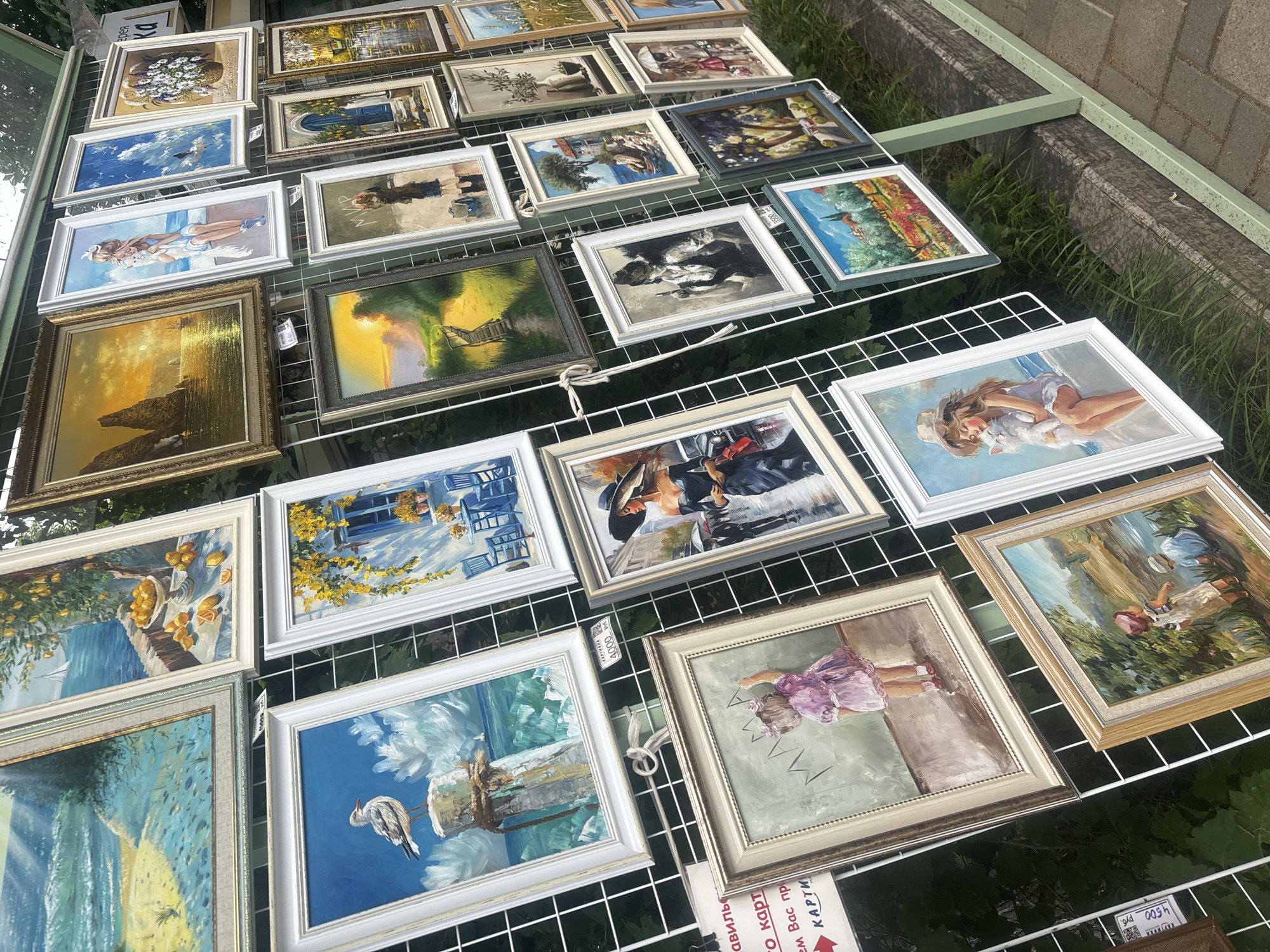 An outdoor art exhibition features a variety of framed paintings displayed on a white grid structure. The paintings depict various subjects, including seascapes, still lifes, portraits, and landscapes. In the foreground, several paintings are visible, including one with a seagull on a post, another of a little girl with "Mama" written above her, and a third showing two children by a body of water. In the background, more paintings are hung, showcasing a sunset over rocky cliffs, a woman with a cat, and a street scene with a woman in a black dress. The exhibition appears to be set up on a sidewalk or paved area with greenery and grass visible in the background.