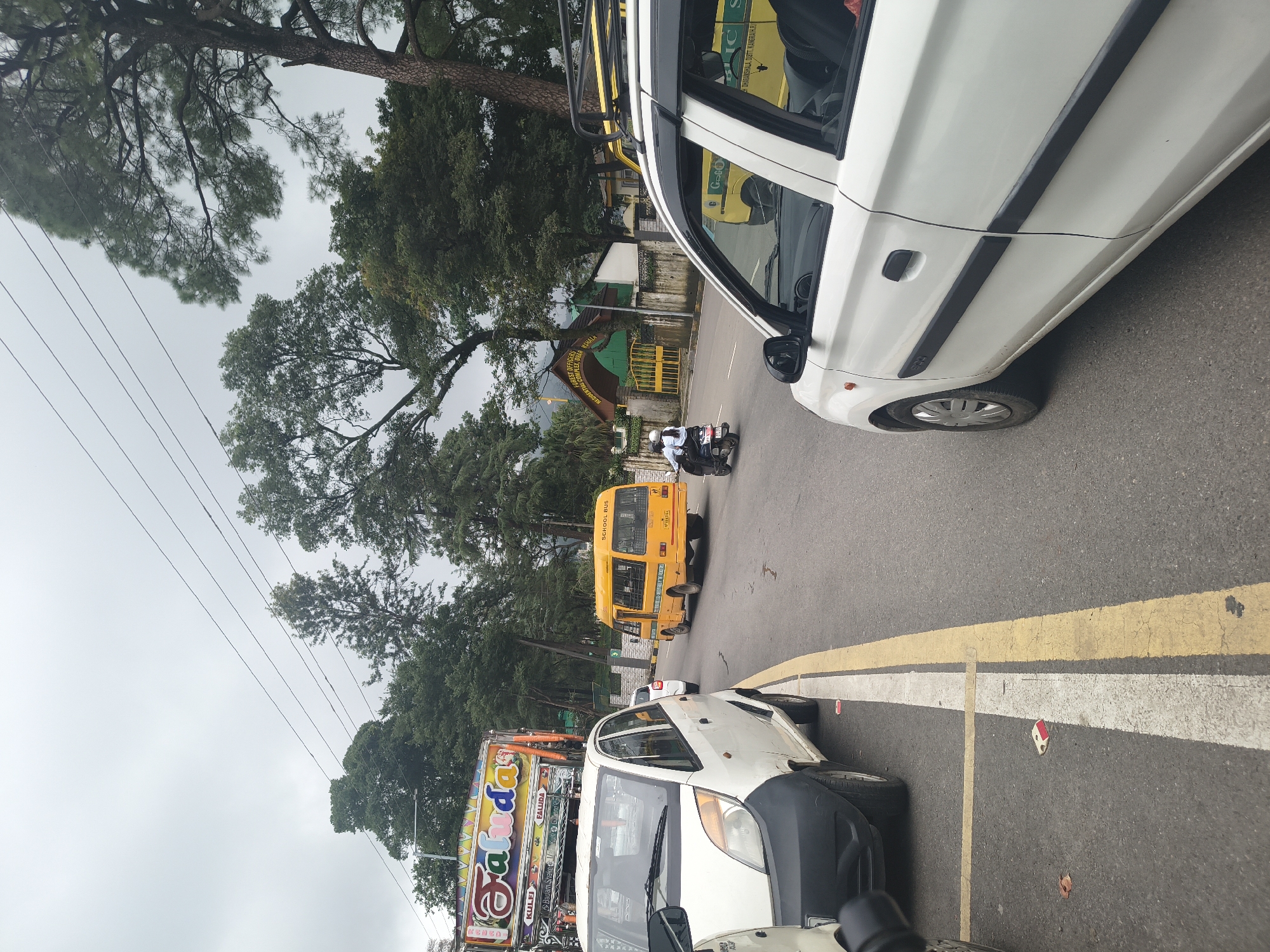 The image captures a street scene in Dharamshala, India, under an overcast sky. In the foreground, a white car is parked on the left side of the road. To the right, another white car, likely a Maruti Suzuki Alto, is in motion, its side door visible. A yellow school bus is traveling down the road in the same direction as the white car. A person on a scooter is also visible, moving ahead of the school bus.
In the background, a truck with colorful signage, including "Falu da" and "Kulfi," is parked. Further down the road, a green and yellow gate is visible, leading to a building with the sign "FOREST OFFICERS RESIDENTIAL COMPLEX, DHARAMSHALA." Trees line the street and the sky is cloudy. The road has clear lane markings, including a white line and a yellow line separating traffic lanes. Some litter is visible on the side of the road.
