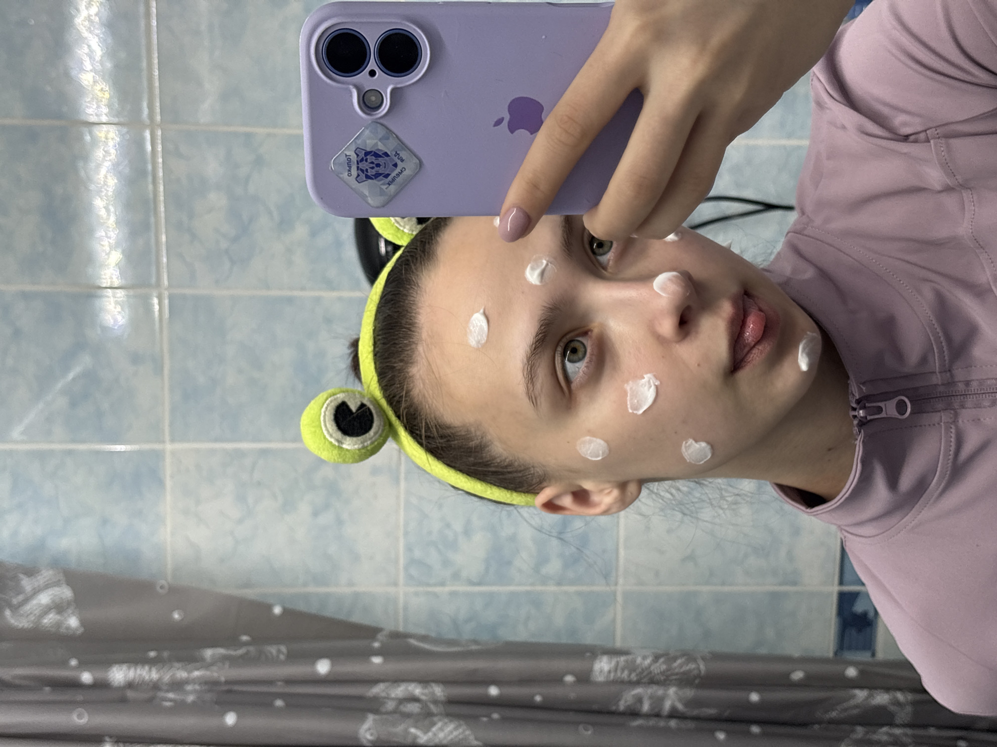 A person takes a selfie in a bathroom. The person is wearing a light purple zip-up jacket and a green frog-themed headband. White cream or lotion is applied in dots on the person's face. The person is holding a purple iPhone to take the photo. The phone has a sticker on the back of the case and the Apple logo. The background includes light blue tiled walls and a gray and white shower curtain. The exact location in Прикамский, Russia is unknown.