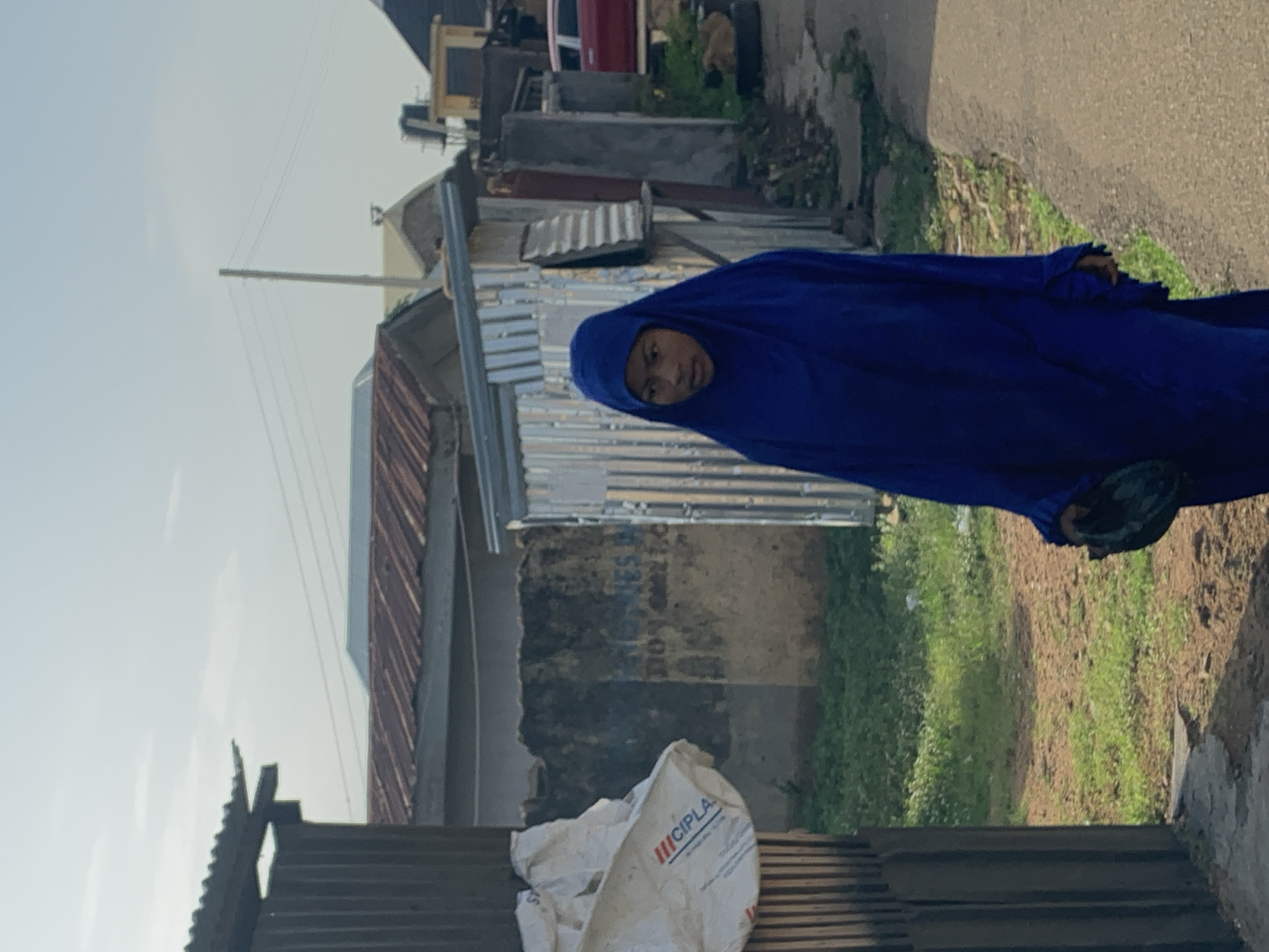 A young person wearing a blue hijab and long blue garment walks on a dirt road in Jos, Nigeria, on a sunny day. The person is holding a dark-colored bag in their right hand. To the left of the person, there is a corrugated metal fence and a white plastic bag with red and blue lettering. Behind the person, there are several buildings with corrugated metal roofs, some in disrepair. There is also a red car parked further down the road. The ground is a mixture of dirt and green grass. The sky is clear and blue.