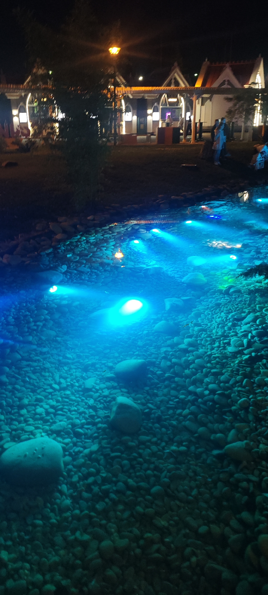 The image captures a nighttime scene in Sirius, Russia, featuring a pond or water feature with illuminated blue lights. The water is shallow, revealing a bed of pebbles and larger rocks. Several circular lights emit a strong blue glow within the water, creating a vibrant effect. In the background, a building is visible, illuminated with warm lights. People are present near the building. A streetlight is visible.