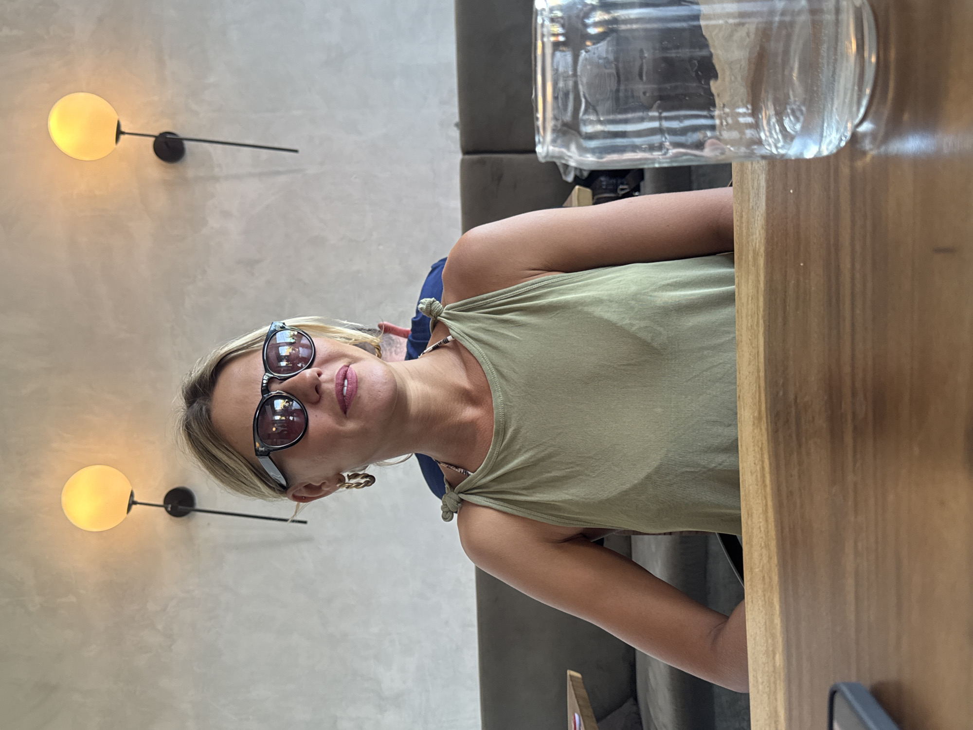A woman wearing sunglasses and an olive-green top sits at a wooden table. A glass of water sits on the table. The background includes a wall with sconce lighting and a gray couch. The photo appears to have been taken indoors. While there is no explicit evidence of the location being in Kuta Selatan, Indonesia, nothing in the image precludes the possibility.