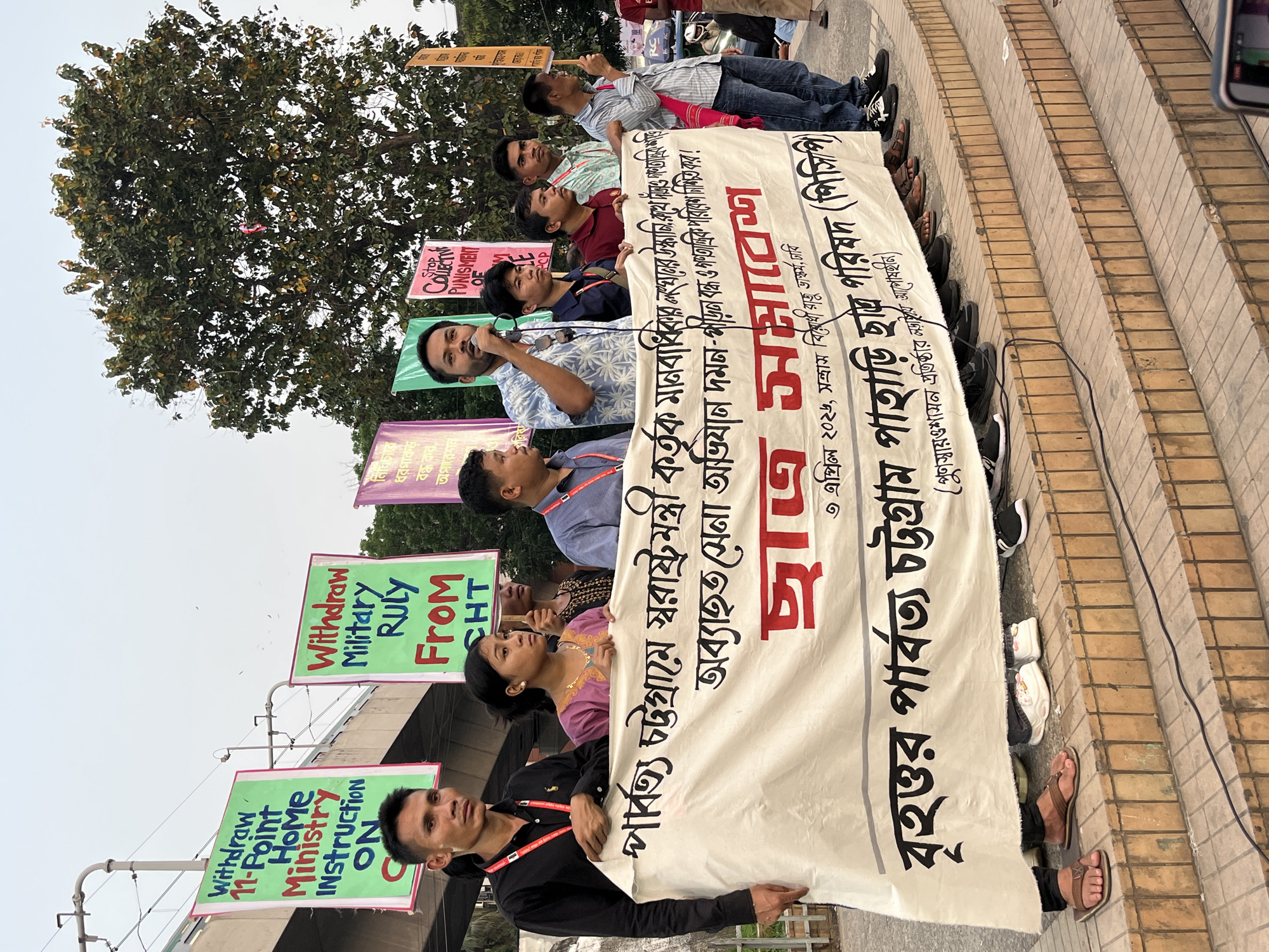 A group of people are protesting in what appears to be Dhaka, Bangladesh. They stand on a staircase, holding signs and a large banner. One sign reads "Withdraw 11-Point Home Ministry Instruction ON". Another sign reads "Withdraw Military RULY FROM CHT". The banner has text in Bengali, including "বৃহত্তর পার্বত্য চট্টগ্রাম পাহাড়ি ছাত্র পরিষদ (পিসিপি)". Several individuals are speaking into microphones. The protest occurs near a road and beneath some trees. The sky is overcast.