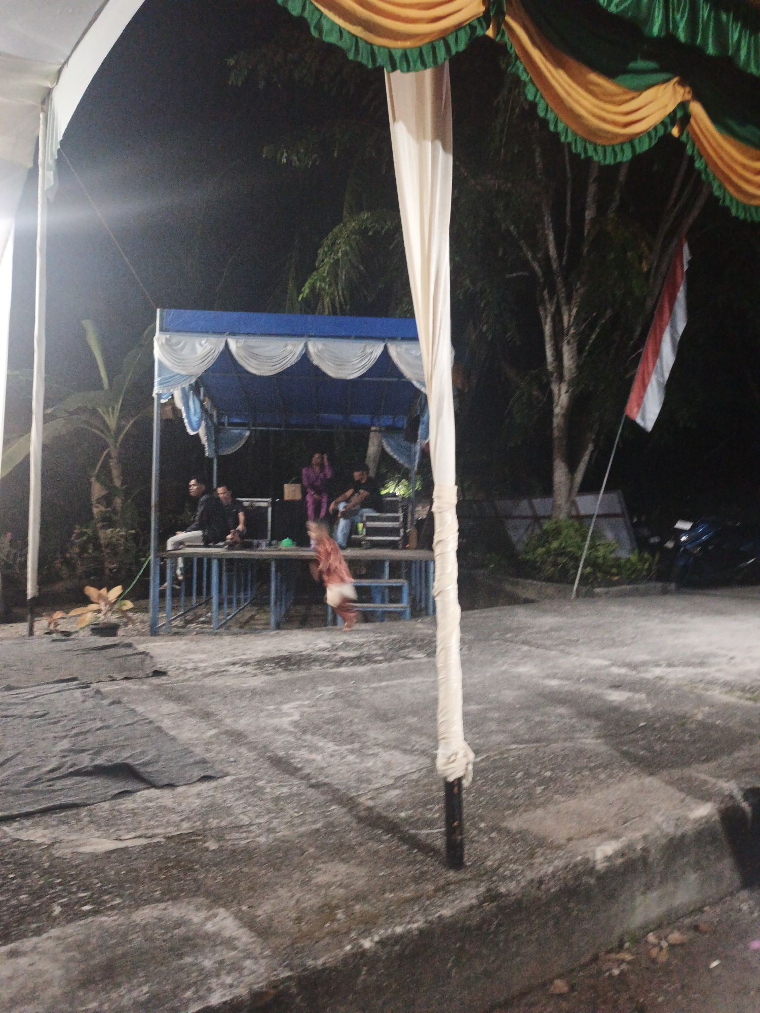 This is a night-time, outdoor photograph taken at a public event, likely a performance or gathering, in Pematang Duku, Indonesia. In the center of the image, a blue and white tent structure is set up on a raised platform, serving as a stage. Several individuals are visible on and around the stage. Two men in dark clothing are seated on the platform to the left, and another man is seated behind them. To the right of the stage, a person in a purple outfit is standing. A young child in a reddish-brown garment is in motion, appearing to be running or dancing in front of the stage, slightly to the right of the frame's center.
In the background, trees and foliage are visible, with the darkness of night suggesting the absence of significant ambient light. A red and white flag, likely the Indonesian flag, is prominently displayed on a flagpole to the right of the stage. In the far right of the background, a motorcycle is parked.
The foreground consists of a paved or concrete surface, with some dark tarp-like material laid out on the left side. A white pole, wrapped in light-colored material, stands prominently in the middle ground to the left, obscuring a portion of the stage and the child. The overall scene depicts a lively community event taking place after dark, with a decorated stage and participants.