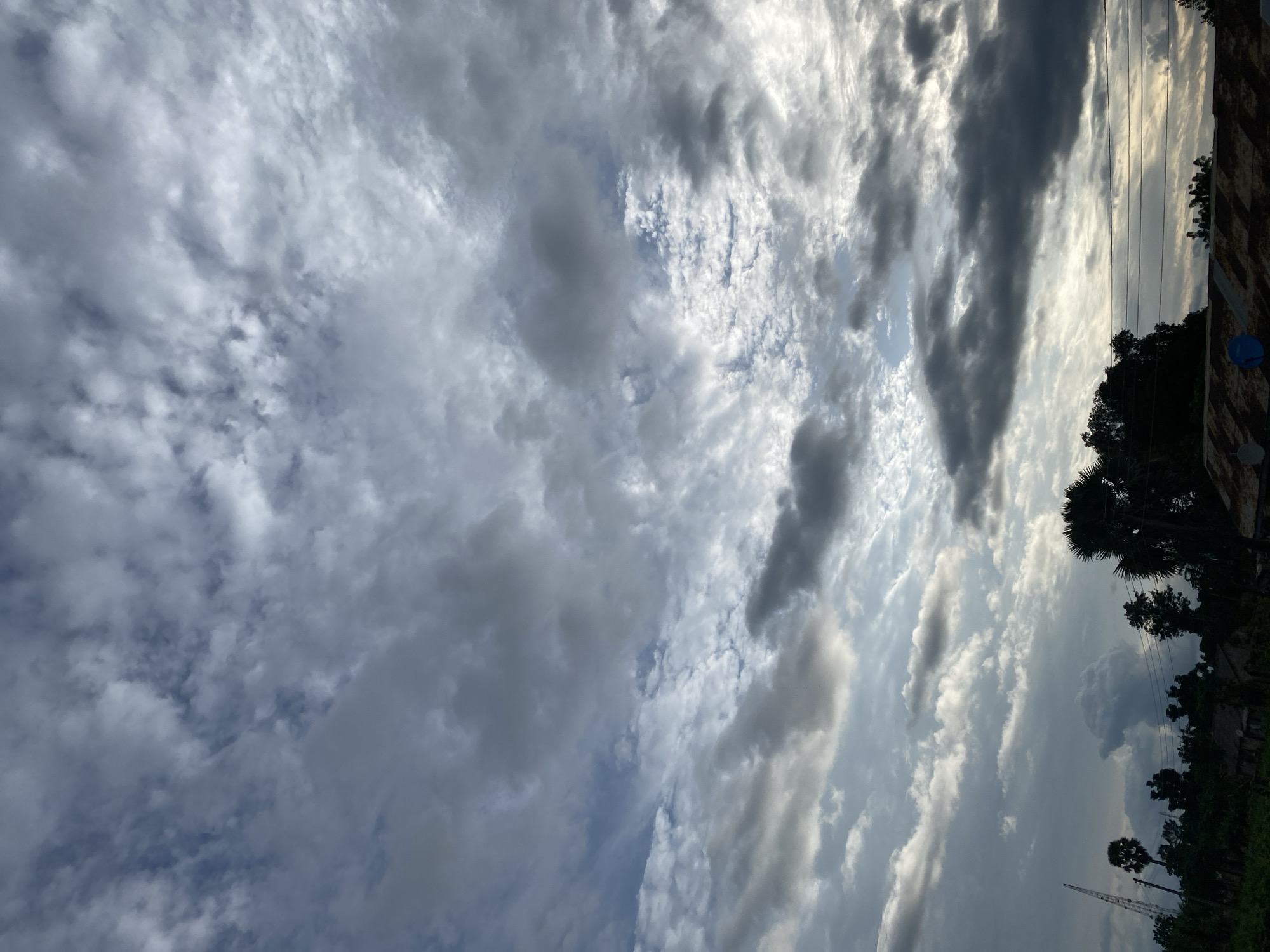 A low-angle, vertical shot captures a dramatic sky filled with a variety of clouds. The sky is a mix of light and dark grays and blues, with patches of bright white where sunlight breaks through. Some clouds are thin and wispy, while others are thick and ominous. In the lower portion of the image, a dark silhouette of trees and buildings is visible, along with utility lines stretching across the frame. A tall, slender tower is also present on the left side. The overall impression is of a powerful and dynamic sky over a landscape. The scene appears to be set in Maigizo, Nigeria, as indicated by the presence of palm trees and vegetation typical of the region, as well as the style of some of the structures. There are no people visible in the image.