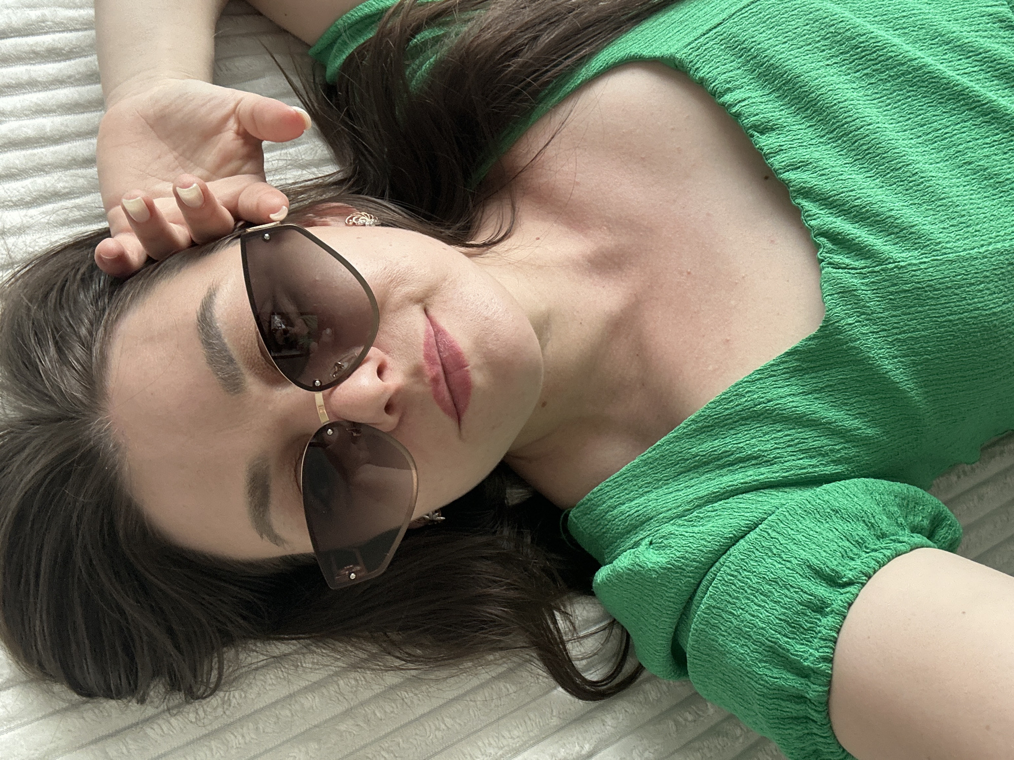 A woman is lying on a white textured surface, presumably a bed, wearing a green off-shoulder top. She is wearing sunglasses with brown tinted lenses and gold frames. Her hair is dark brown. Her left hand rests on her forehead, fingers slightly curved. Her lips are visible, painted with a shade of pink. An earring is visible on her left ear. The location is unspecified and therefore can not be identified as being in Chaikovsky, Russia, or any other location.