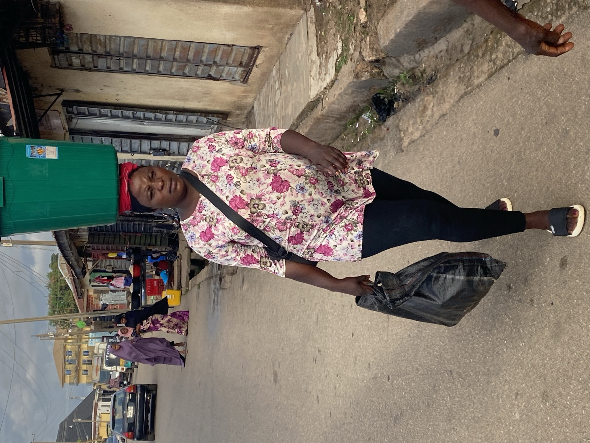 A woman walks down a paved street in Jos, Nigeria, carrying a large green bucket balanced on her head and a black plastic bag in her right hand. She wears a floral print three-quarter sleeve top with pink and white flowers, black leggings, and black and white sandals. A black shoulder bag is slung across her body. Her expression is neutral. To her left, a black sedan is parked facing away, and a "keke napep" (motorized tricycle) is visible further down the road. In the background on the left, a multi-story yellow building stands under a cloudy sky with visible utility lines. To her right, along the roadside, two women in traditional attire walk past market stalls displaying goods like sacks of grain and a yellow plastic container. One woman wears a long purple garment, and the other a patterned dress with a black head covering, appearing to hold a child. The market stalls feature dilapidated wooden or corrugated metal shutters. A concrete drainage ditch runs alongside the road. The hand and arm of a third person are visible in the lower right corner.