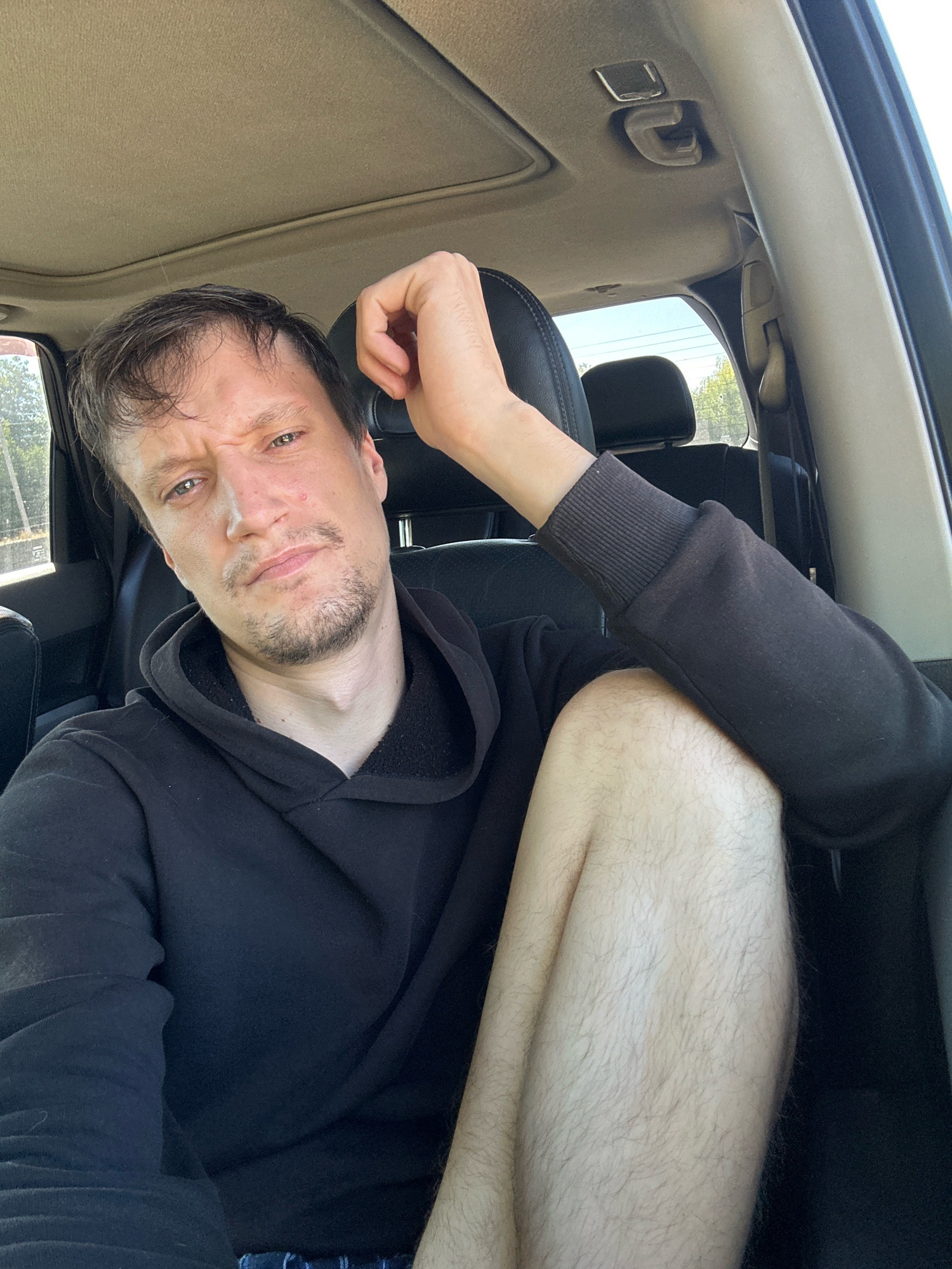A man is sitting in the driver's seat of a car, looking at the camera with a skeptical expression. His left arm is resting on the back of the seat, and his right leg is bent and crossed over his left. He is wearing a black hoodie. The interior of the car is visible, including the headliner, seats, and a portion of the dashboard. The scene appears to be outdoors, with sunlight filtering into the car. The image does not provide any specific details or context that would allow for the identification of the location within the city of Makiv, Ukraine.