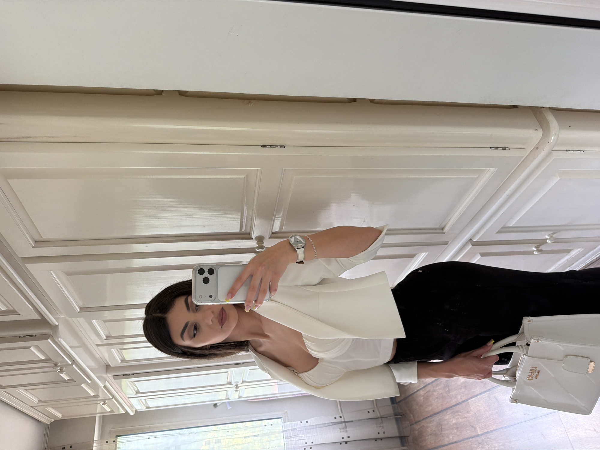 A young woman is taking a selfie in a room with white paneled walls and cabinets. She's holding her phone in her left hand, and wearing a white blazer over a light-colored top, and black pants. In her right hand, she's holding a white handbag. It's impossible to determine if this is in Gabrovo, Bulgaria, from the image.