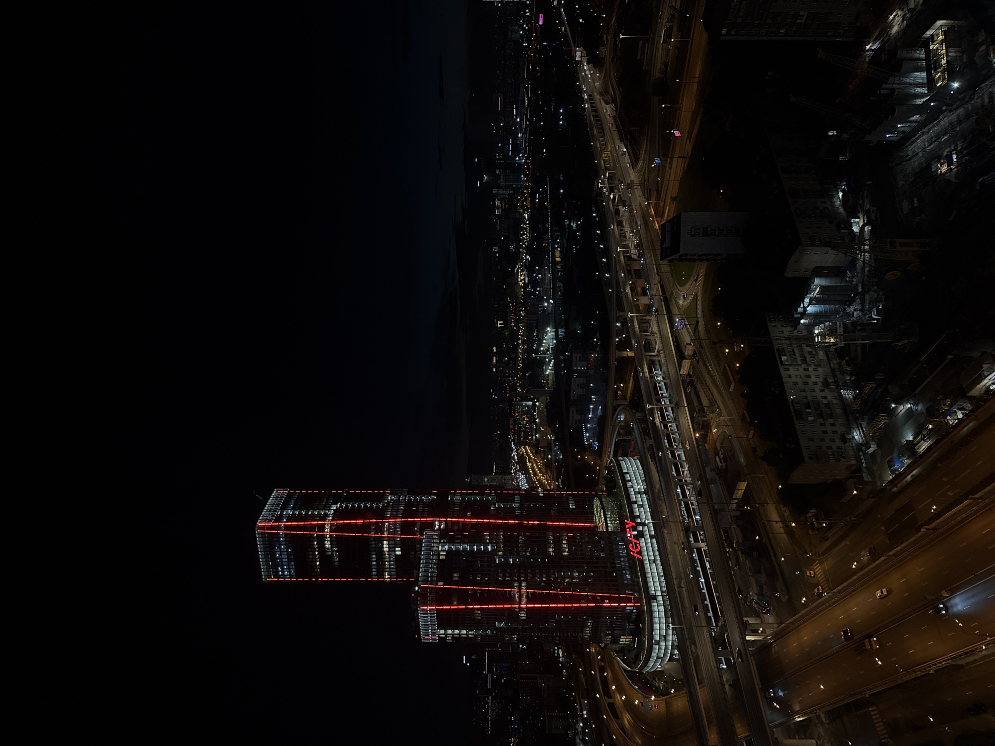 An aerial, nighttime view of the city of Khimki, Russia, showcases the IQ-City business center, a prominent modern skyscraper complex. The IQ-City buildings are illuminated with vibrant red vertical lights, creating a striking contrast against the dark sky. The complex is surrounded by a network of multi-layered highways and overpasses, with cars visible on the roads. In the foreground, several lanes of traffic move along illuminated highways. The background reveals a sprawling cityscape with numerous lights from other buildings and streetlights, extending into the distance. Construction sites with cranes and heavy machinery are also visible in the lower right portion of the image, indicating ongoing development in the area. The overall scene depicts a busy urban environment at night, dominated by the illuminated architecture and infrastructure.