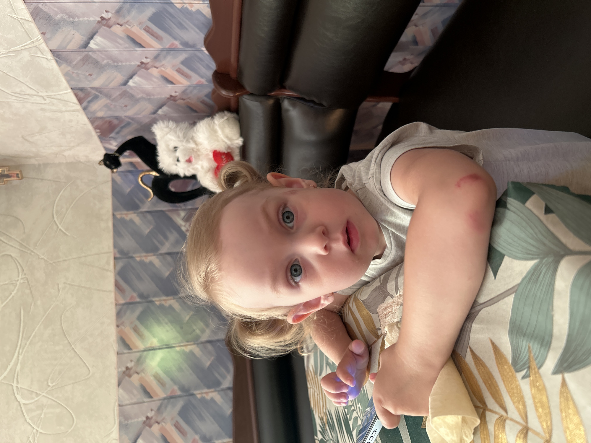 A young child with blonde hair in pigtails and blue eyes is leaning on a table covered with a tablecloth featuring a floral pattern of green and gold leaves. The child is wearing a gray t-shirt and has a small abrasion on their left elbow. In the background, a white teddy bear with a red heart is visible, perched on a dark leather booth. Behind the booth, the wall is decorated with a patterned wallpaper. The lighting suggests an indoor setting, possibly a restaurant or cafe in Gelendzhik, Russia.