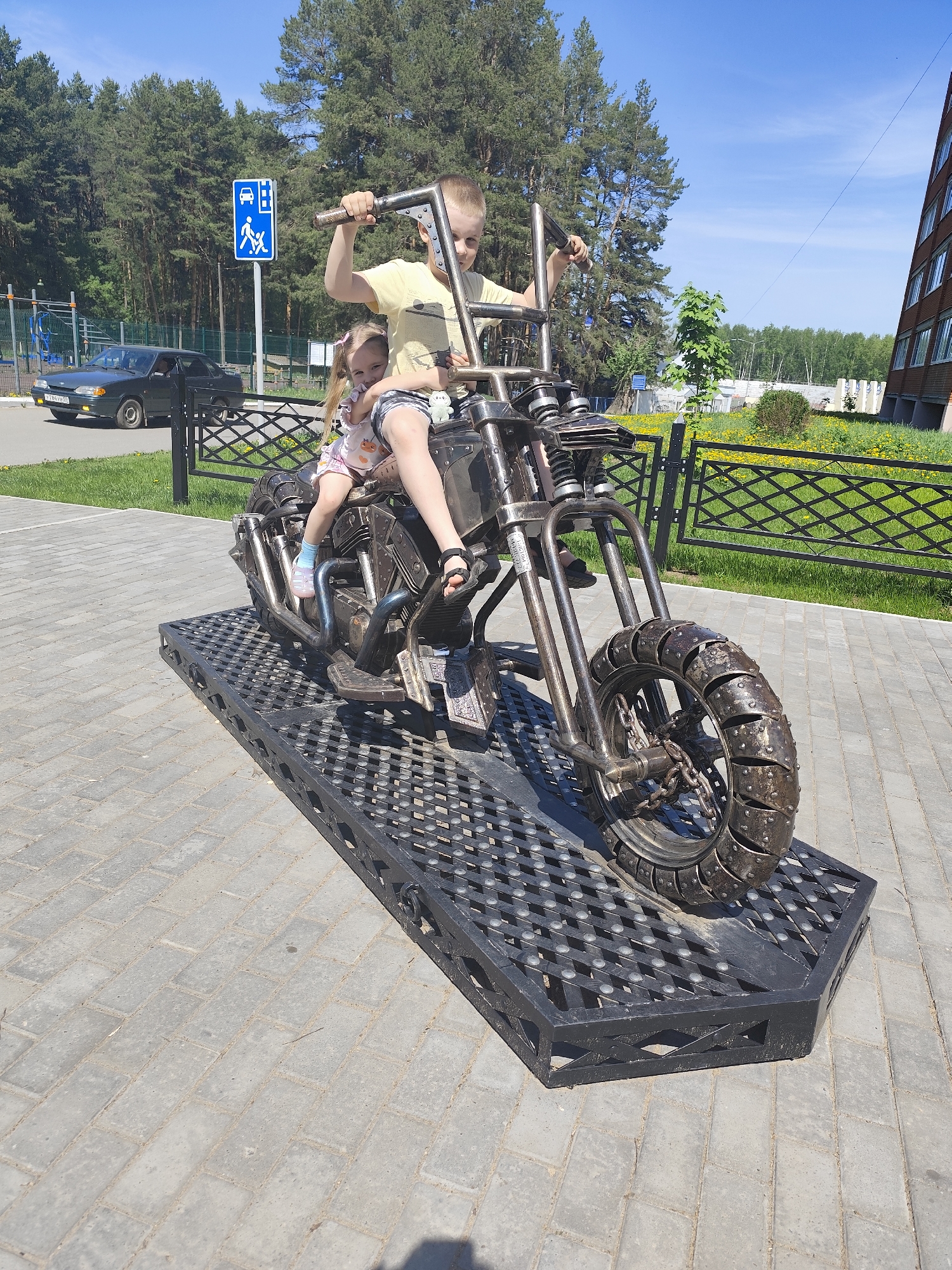 Two children, a boy and a girl, are on a motorcycle sculpture made of metal in Chaikovsky, Russia. The boy, wearing a yellow shirt and patterned shorts, is holding the handlebars. The girl, wearing a light-colored dress with a print, is sitting in front of him. The motorcycle sculpture is positioned on a black metal platform, which is on a paved area. In the background, there are trees, a building, a sign with a pedestrian crossing symbol, and a dark-colored car. A black metal fence runs along the background as well. The sky is blue.