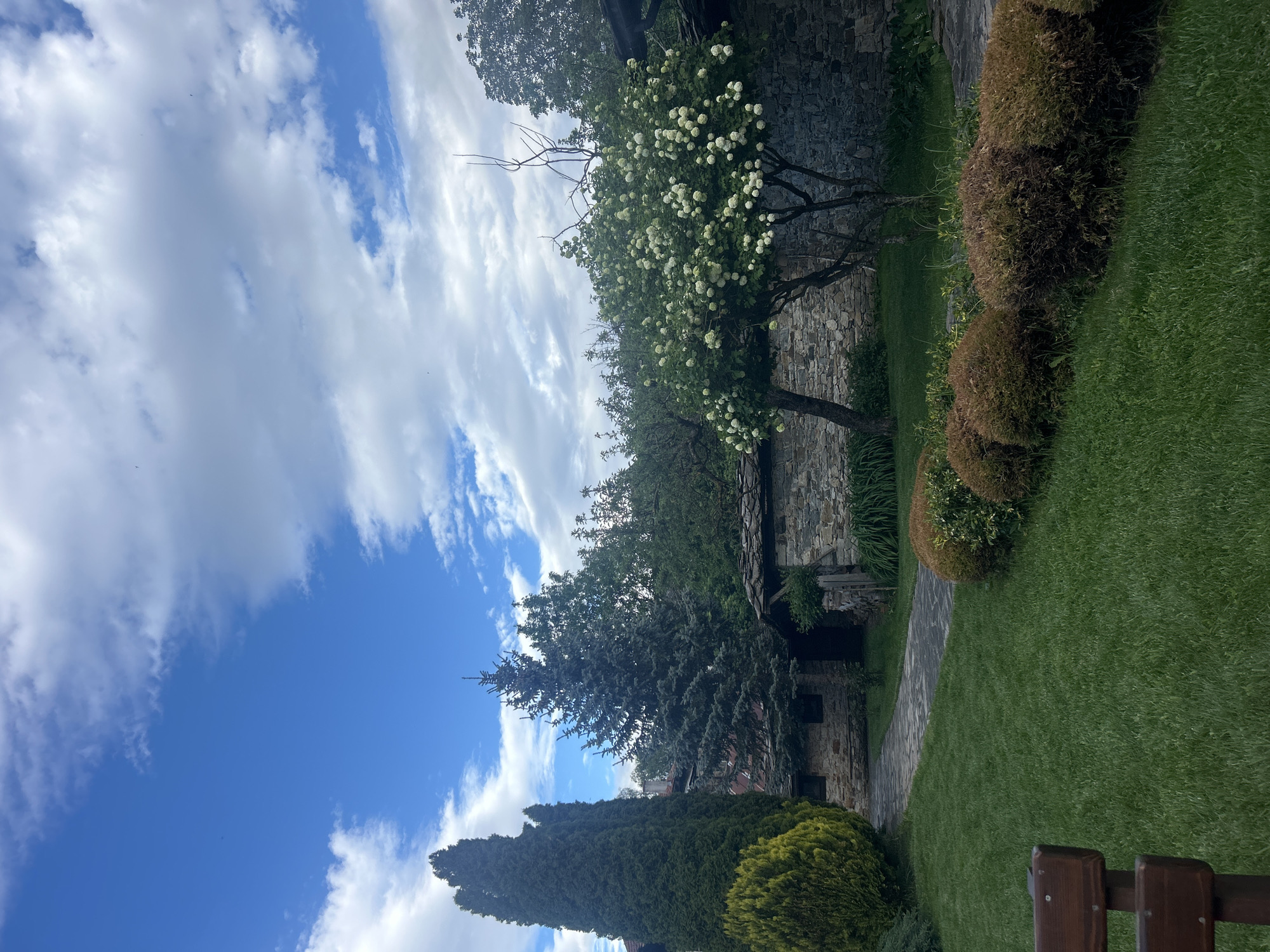 The image shows a stone structure in Dolen, Bulgaria, with a stone wall and building visible behind trees and shrubs. The foreground includes a lawn, a stone path, and manicured bushes. The sky is blue with scattered white clouds. The image is taken from a low angle, slightly tilting upward towards the sky.