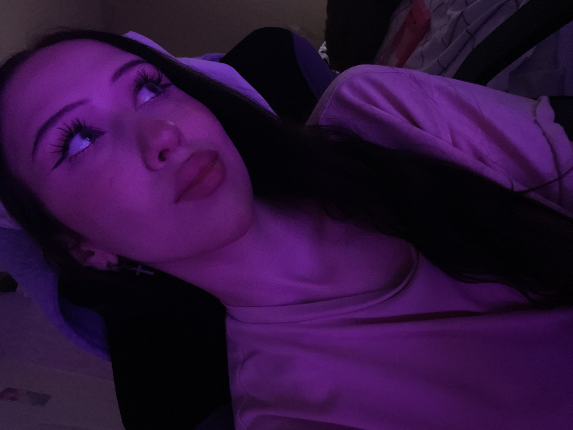 A young woman with long dark hair is shown from the chest up, bathed in purple light. She is wearing a light pink t-shirt. Her head is tilted slightly to the right, and she is looking away from the camera. She has prominent eyeliner and long eyelashes. A cross earring is visible on her left ear. The background is dark, with hints of a chair and bedding. The location is described as the city of Votkinsk, Russia.
