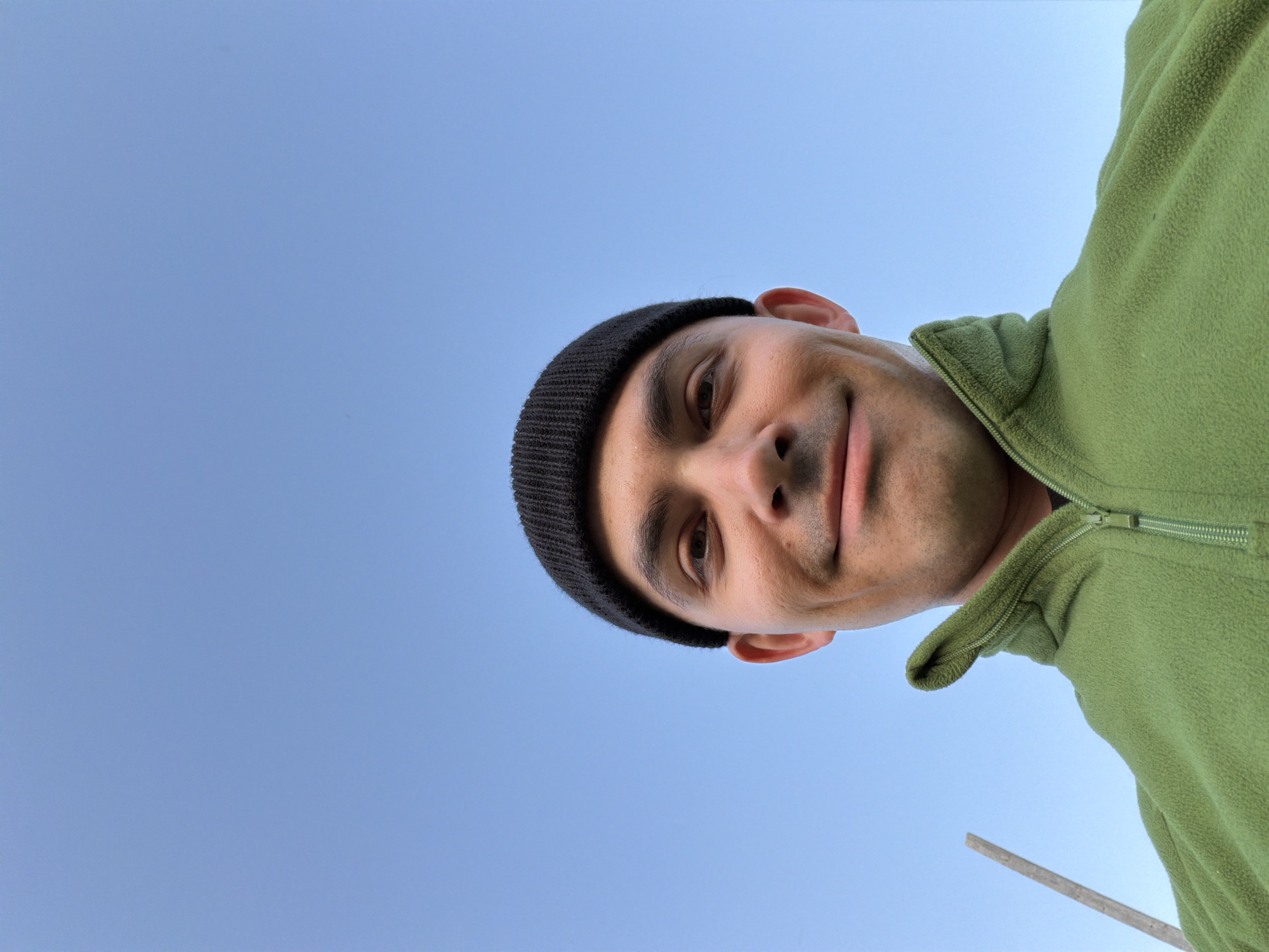The image captures a man's face and upper body, photographed from a low-angle perspective against a bright, clear blue sky. The man is positioned from the left side of the frame, with his head turned slightly upwards and his body extending towards the middle-right. He has light-medium skin, dark eyes, and a slight, closed-mouth smile while looking directly at the camera. He is wearing a black knit beanie and an olive-green fleece jacket with a visible zipper running down the center-right side of the frame. The background is a uniform, cloudless blue sky, indicating a clear and sunny daytime setting outdoors. A slender, light-colored object, possibly a stick or wooden pole, is partially visible at the very bottom-right corner. No text is visible in the image. The scene takes place outdoors, in the context of Kechur, Russia.