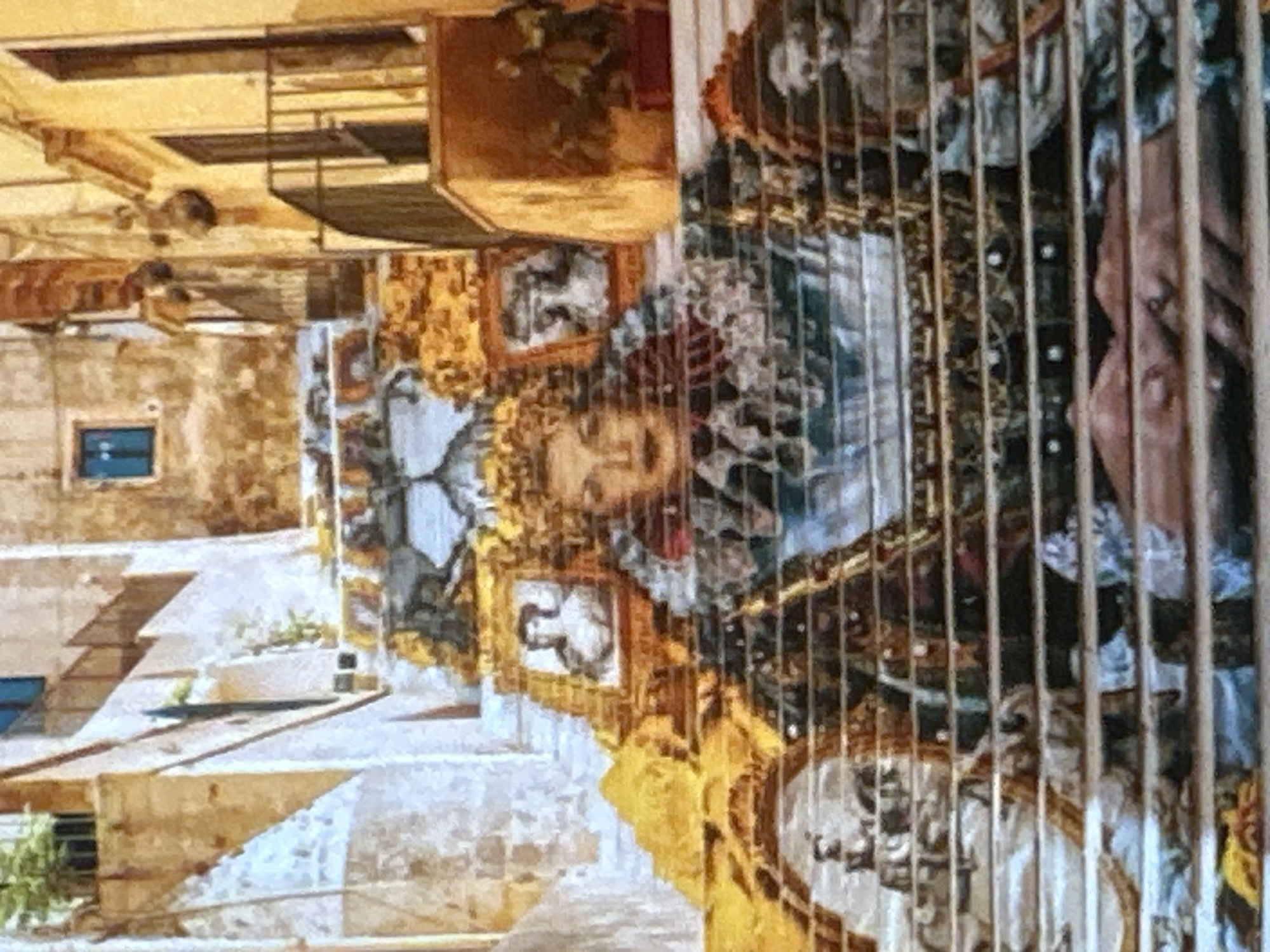 The image depicts a long, wide staircase decorated with a colorful mural that extends across its risers. The mural features several portraits, including a prominent central portrait of a woman with fair skin and dark hair, adorned with elaborate clothing and jewelry. Smaller portraits of individuals are interspersed throughout the design, framed in ornate gold borders. The staircase is set against a backdrop of aged buildings with yellow and tan facades, some featuring balconies and windows. The overall scene suggests an outdoor urban environment, likely a public space within a historic district. The mural appears to be painted or printed directly onto the stair treads, creating a vibrant and artistic transformation of the architectural element. The lighting in the image suggests daytime, with shadows cast on the buildings and stairs.