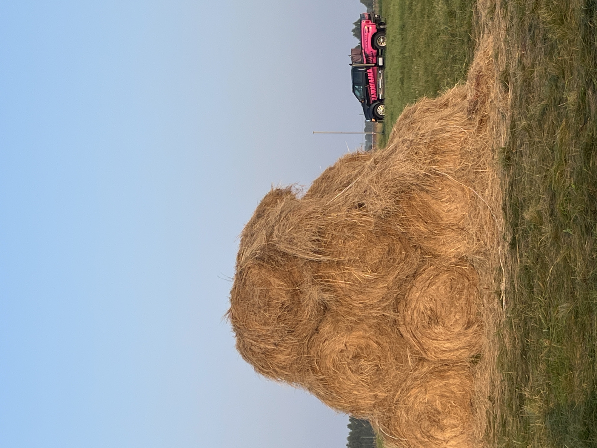 In Surakovo, Russia, a large haystack is situated in a grassy field under a clear blue sky. To the right of the haystack, a black pickup truck with pink accents and the Cyrillic text "ЗАЖИГАЛКА" (Zazhigalka, meaning "lighter" or "spark plug") on its side is parked on the grass. The truck has a custom pink bed. Trees line the horizon in the distance.