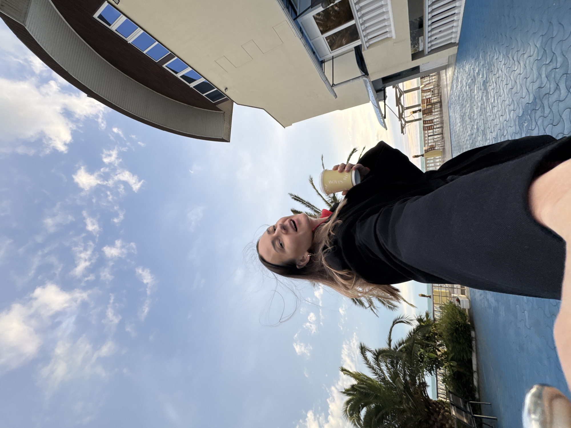 A woman is visible in an upward-angled outdoor shot in Sochi, Russia. She is holding a cup. Behind her are palm trees, a patterned blue surface, and buildings. One building has a rounded section with windows. In the distance, there are chairs and tables. The sky is blue with white clouds.