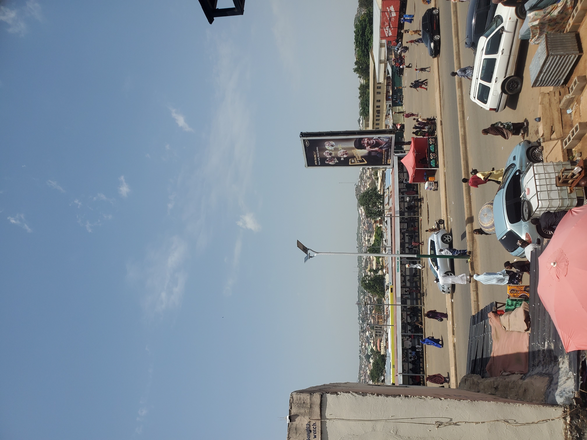 This media file captures a street scene in Jos, Nigeria. The image is oriented vertically, with the sky occupying the left half and the street scene filling the right. Several people are visible, some walking along the street, others standing near parked vehicles. The weather appears to be clear with a bright sky and scattered clouds. The time of day seems to be daytime, possibly late morning or early afternoon, given the lighting.
Objects in the scene include various cars, including a white van and a light blue car. There are also market stalls with visible goods, such as crates of produce and fabric. A prominent billboard displays a man's face and text, suggesting an advertisement. Another advertisement is visible on a building in the background, featuring a yellow and white stripe design. A street lamp with a solar panel is also present. The overall setting is an urban environment with buildings lining the street, some appearing to be shops or residences. The ground is paved, and there are indications of a busy commercial area.
The activity taking place appears to be everyday street life, with people going about their business. The atmosphere seems relatively calm, with no obvious signs of distress or urgency among the individuals. The presence of market stalls and vehicles suggests a functioning urban economy.
Visible text includes "bundles. watch." written on the side of a building in the foreground. The billboard also has some text, but it is too small to be legible. The advertisement on the background building also contains some text, but it is also unreadable.